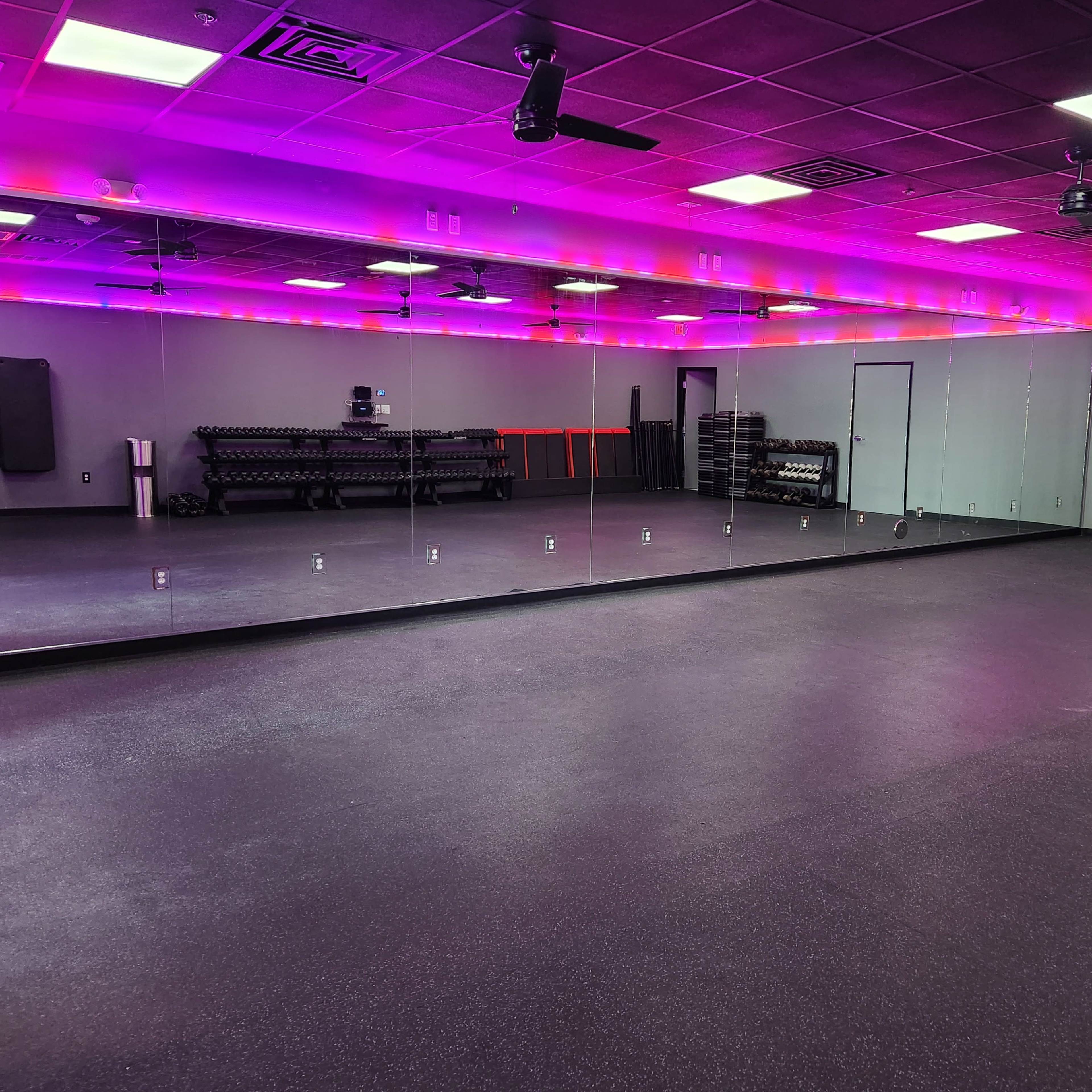 A spacious fitness studio with a large mirrored wall, vibrant lighting, and exercise equipment organized along the sides.