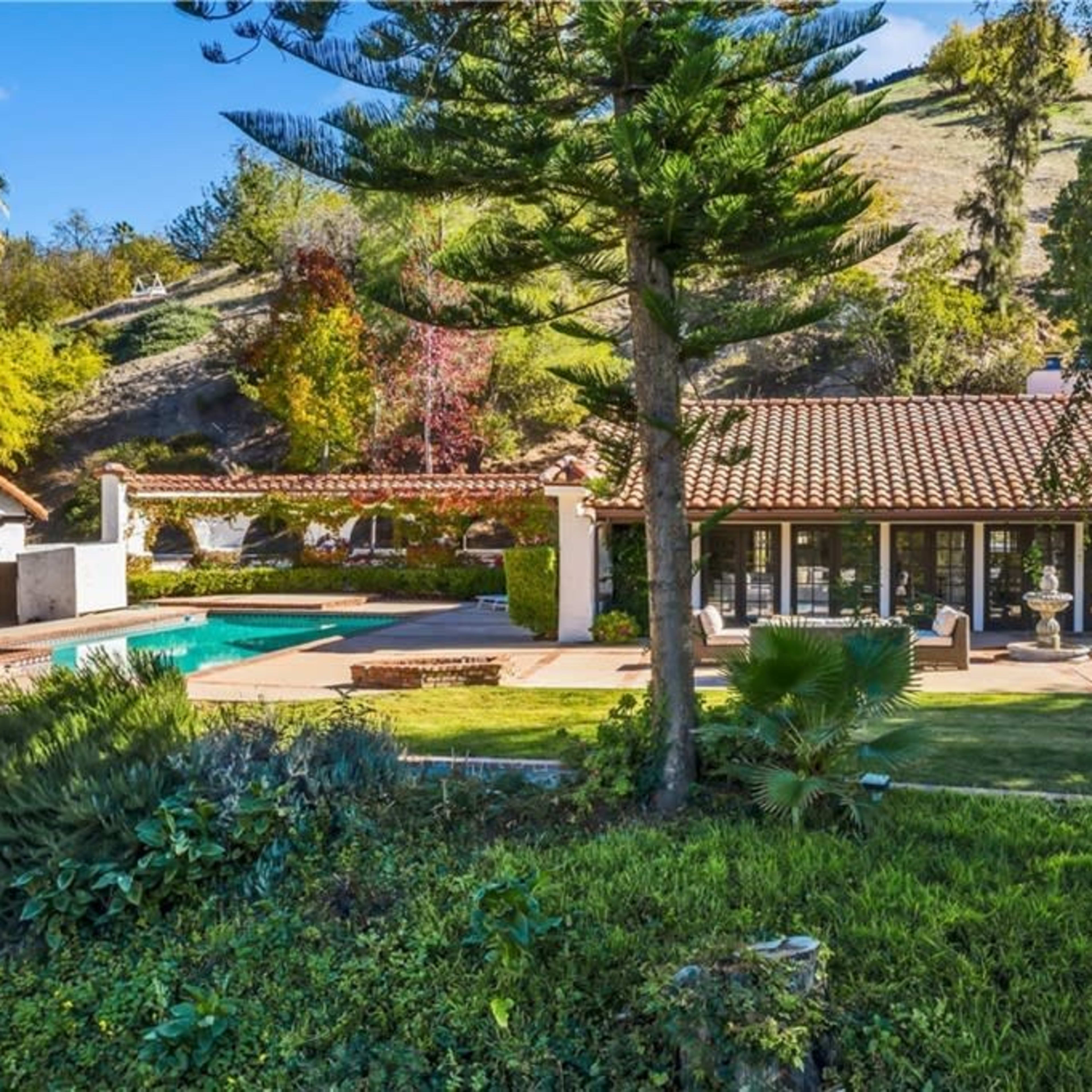 A residential property features a pool surrounded by landscaping, with a hillside in the background and a Spanish-style house adjacent to it.