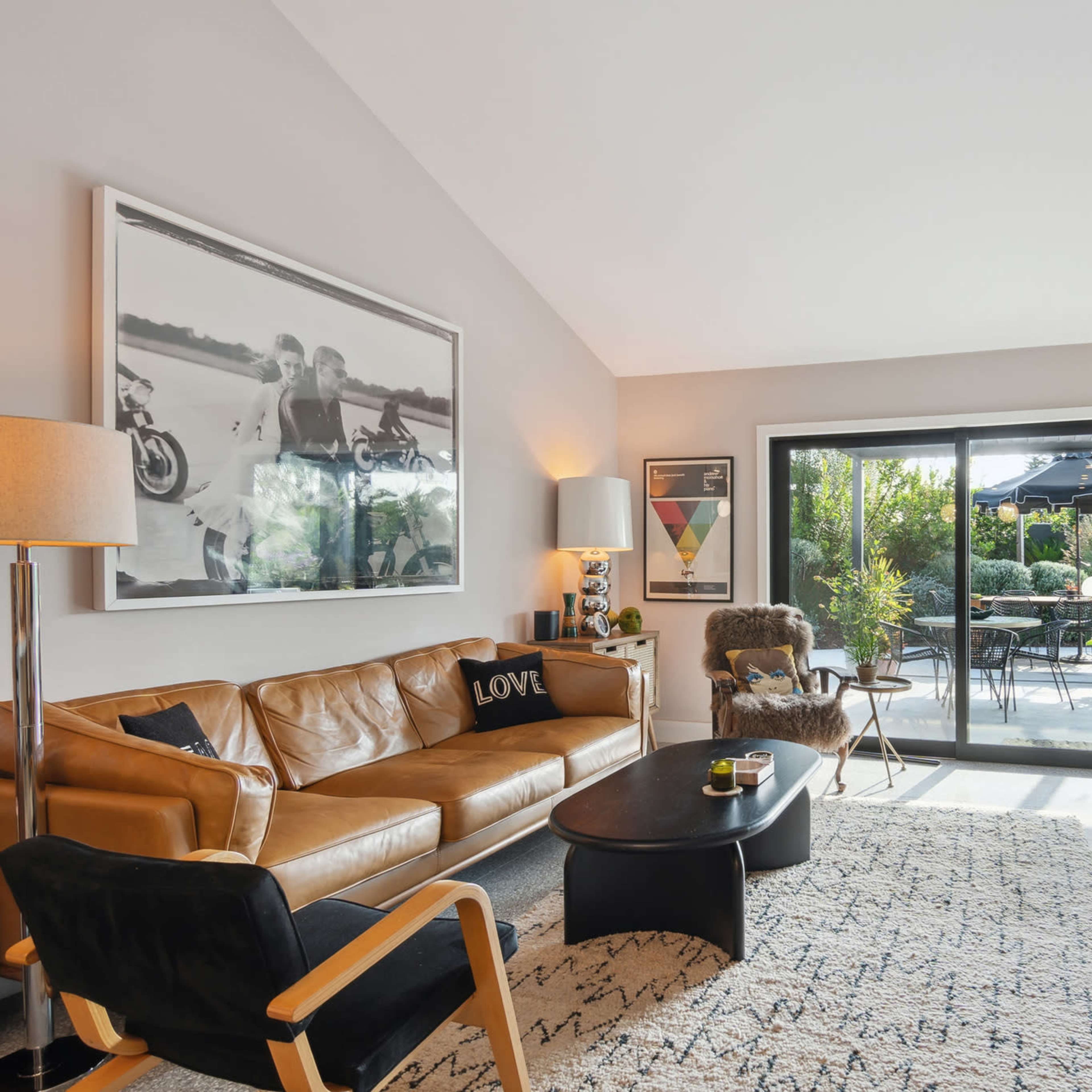 A modern living room with a brown leather sofa, a black coffee table, and large glass doors leading to an outdoor patio area filled with greenery.