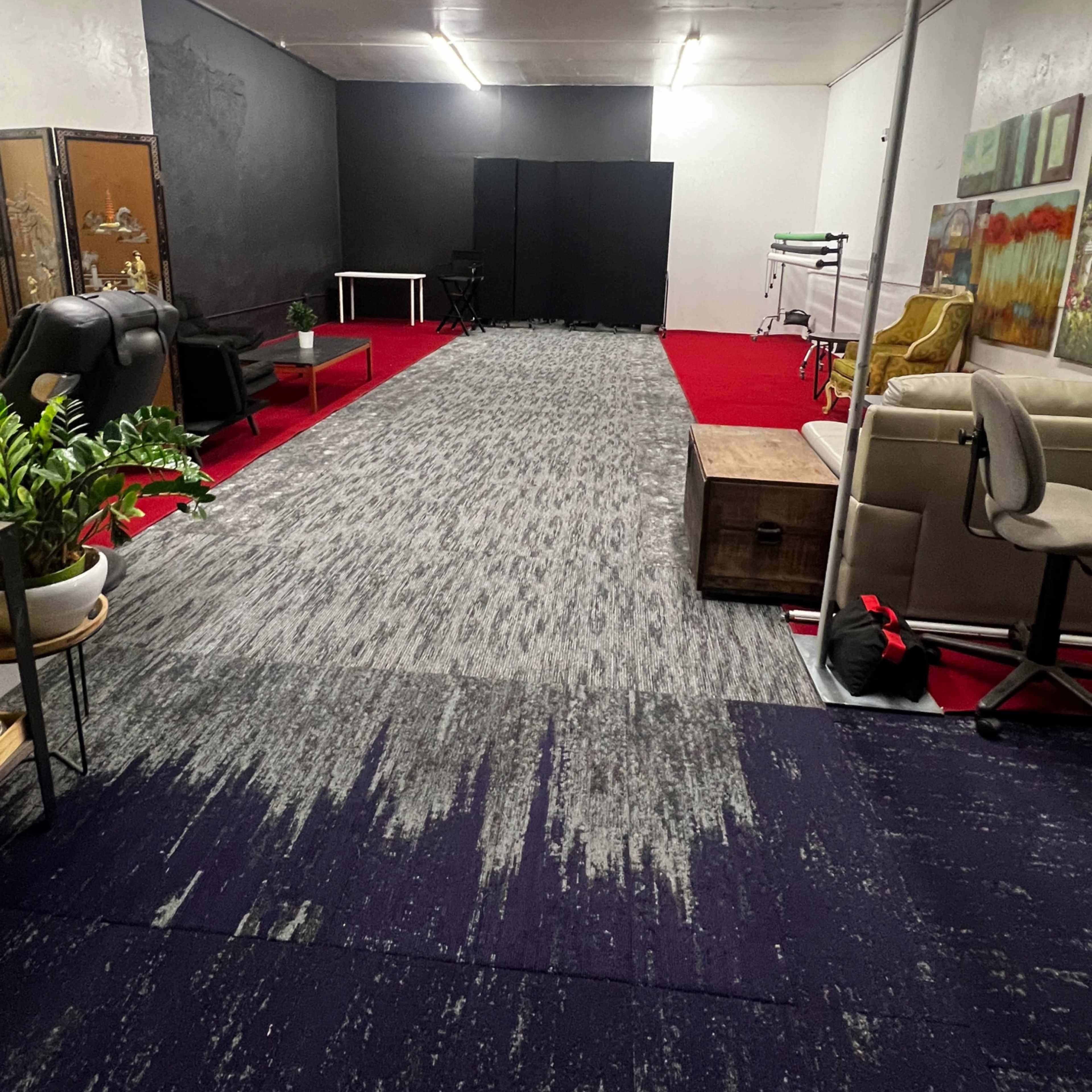 An empty room with red carpet, gray flooring, and various pieces of furniture including chairs and a table along the walls.