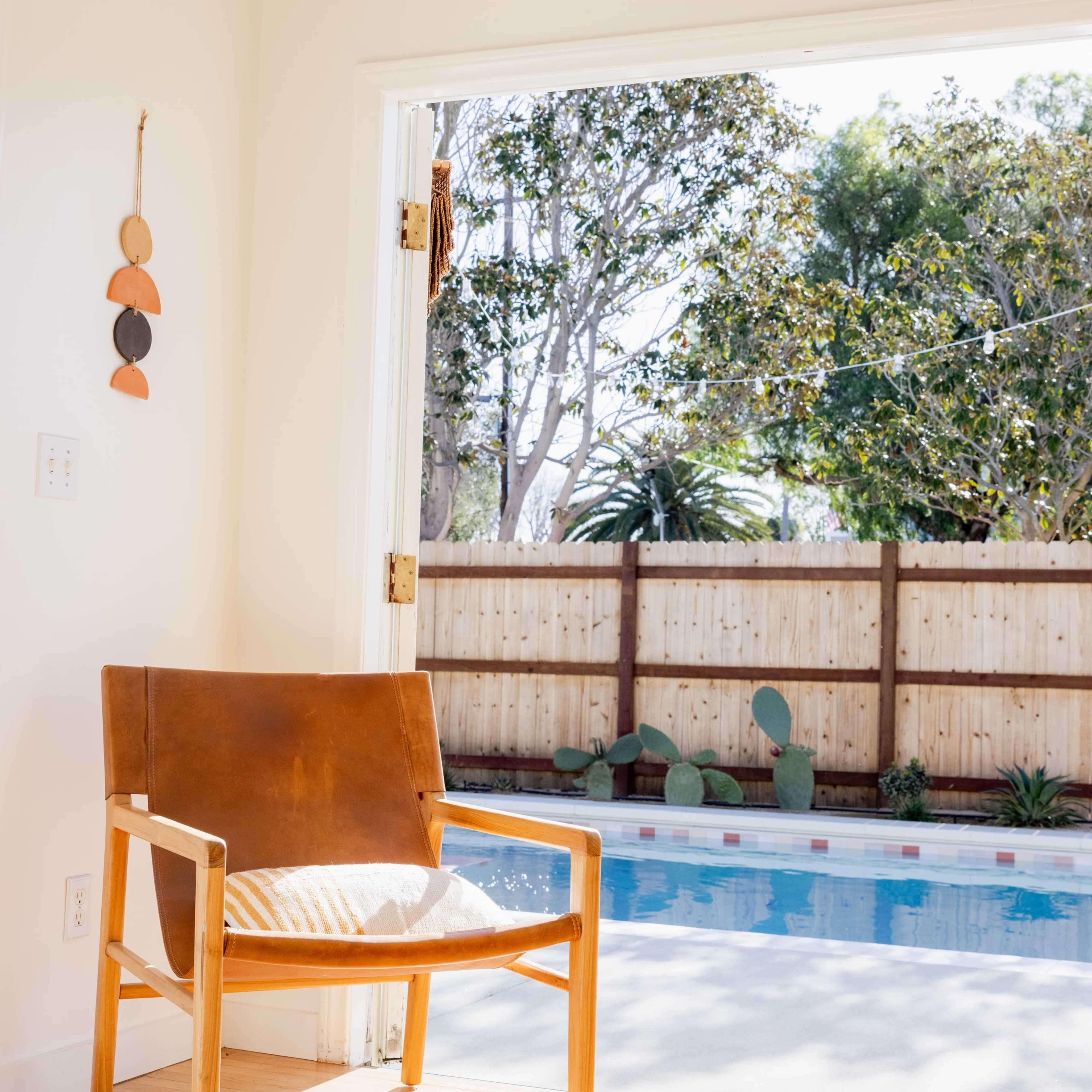 A leather chair sits in a brightly lit room beside an open door leading to a backyard pool area surrounded by trees and a wooden fence.