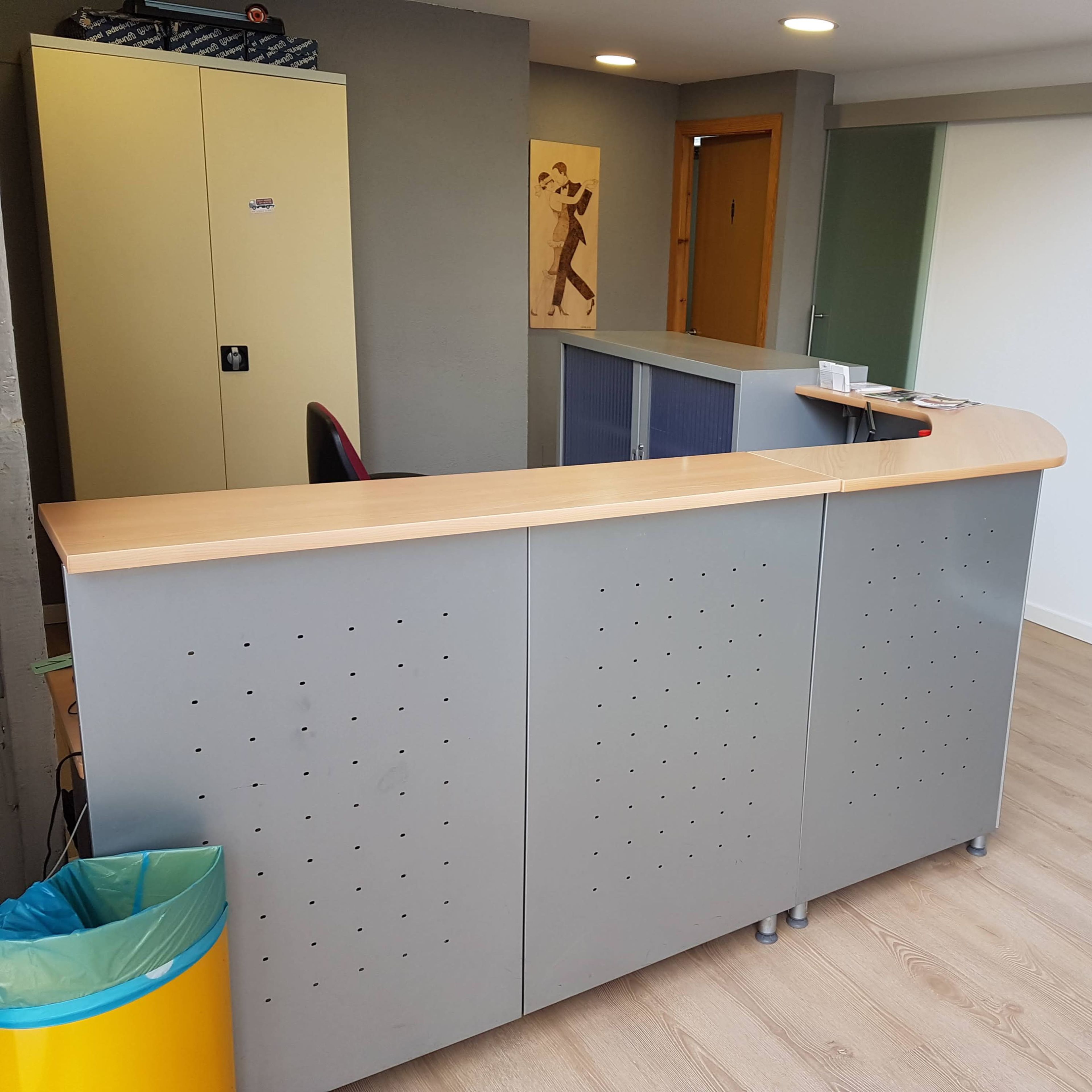 A reception area with a gray desk, a chair, and a trash bin in a well-lit room.