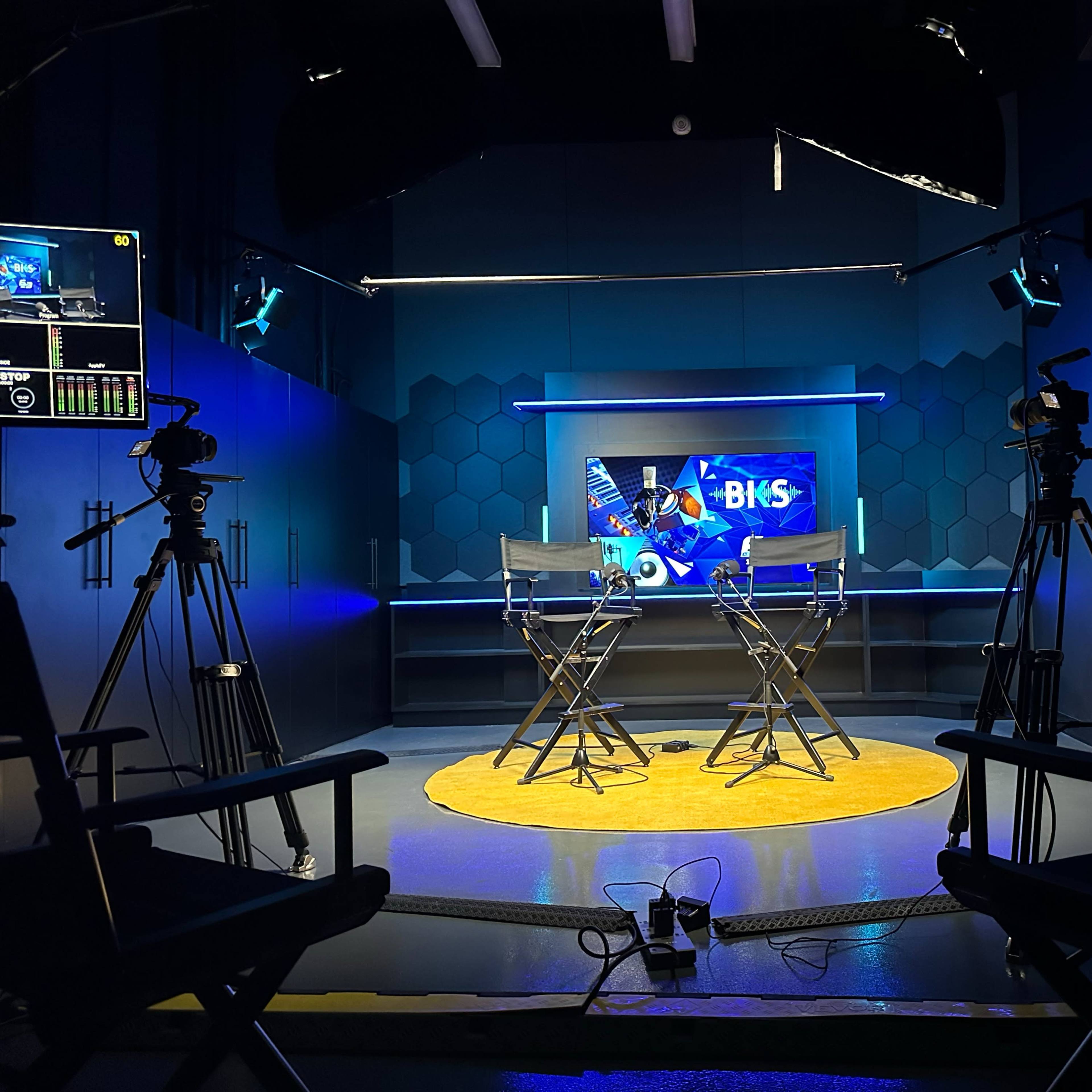 A television studio setup with two director's chairs facing a brightly lit screen, flanked by cameras and staging equipment.