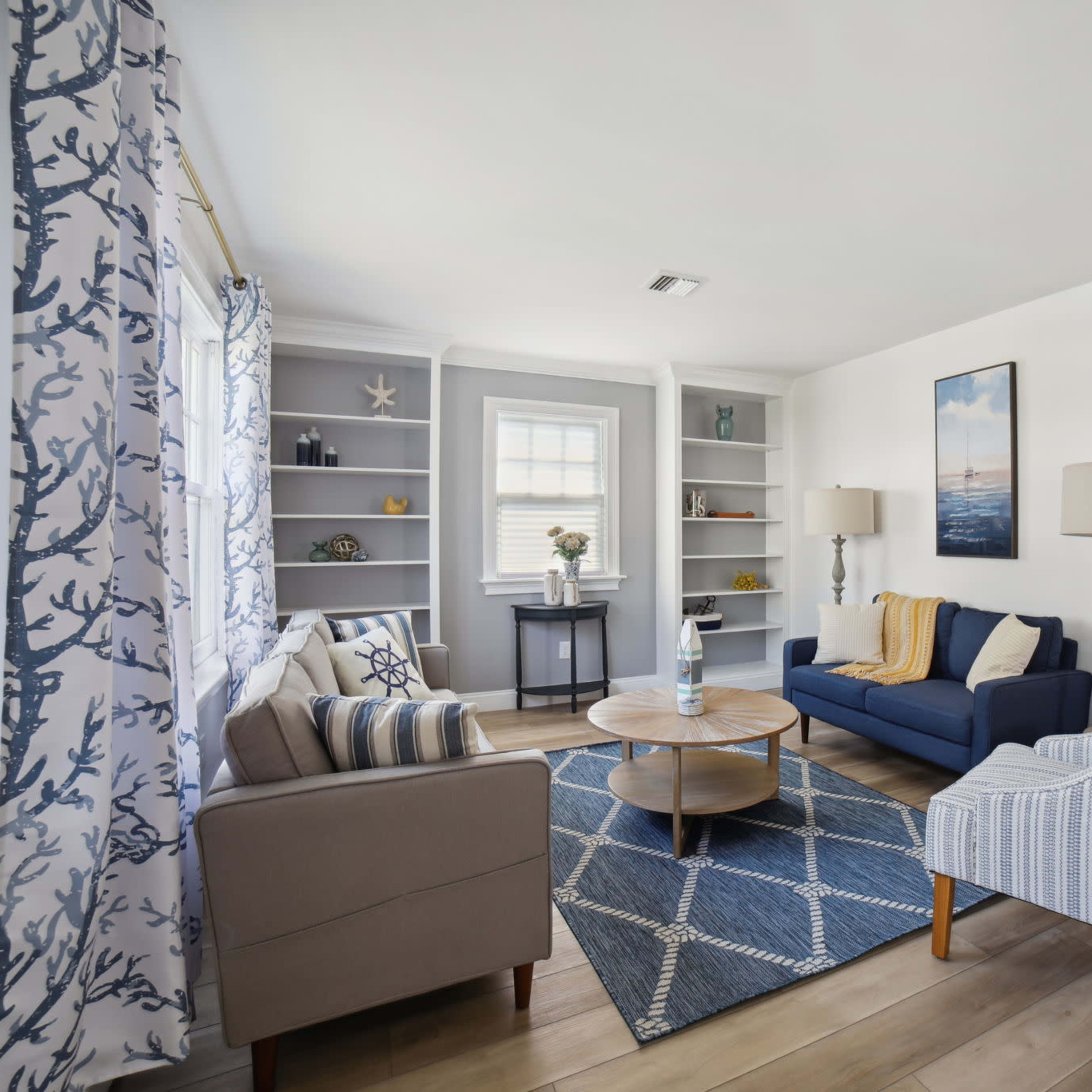 A modern living room with a blue sofa, an armchair, a round coffee table, decorative shelves, and large windows with patterned curtains.