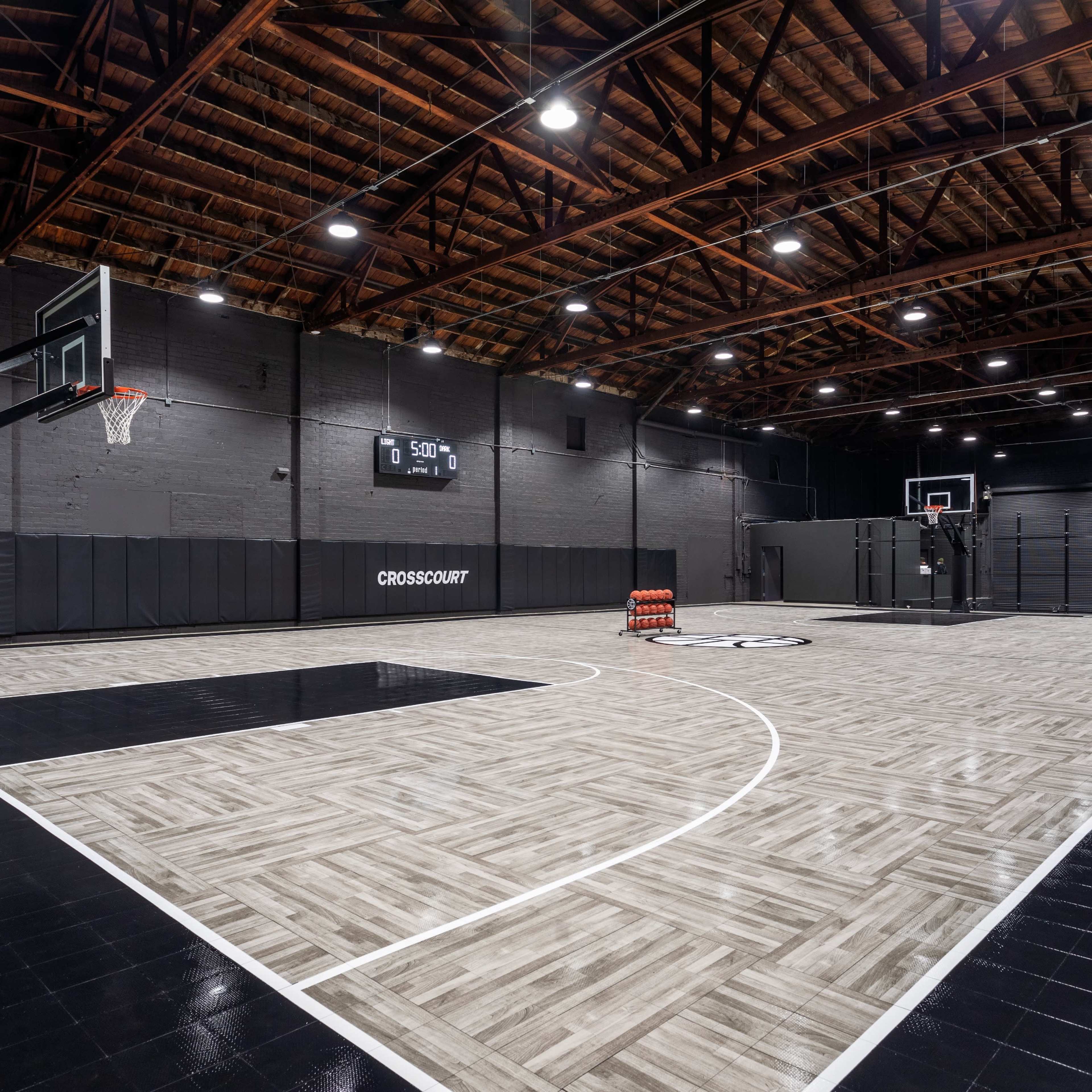 A basketball court featuring wooden flooring, two basketball hoops, and a spacious, well-lit interior with black walls and a high ceiling.
