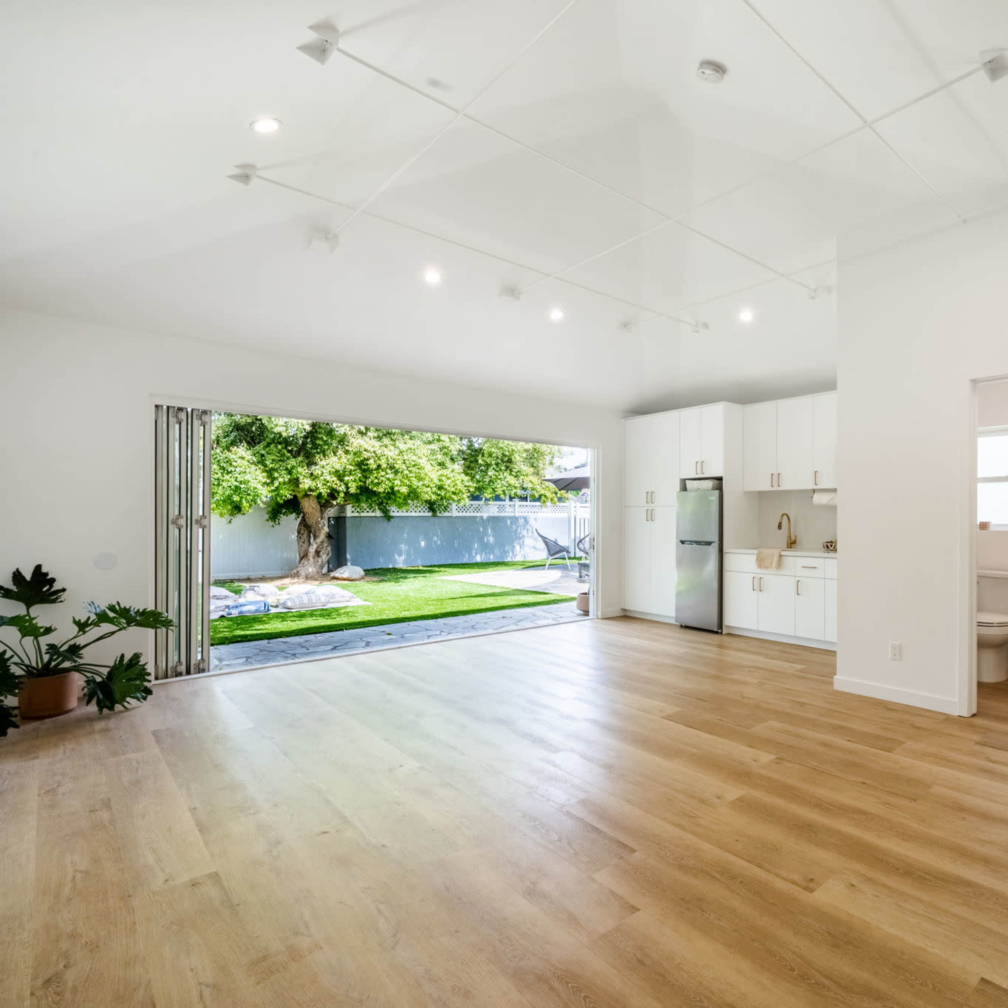 A spacious, well-lit room features an open layout with a view of a backyard, showcasing a kitchenette and a bathroom area.
