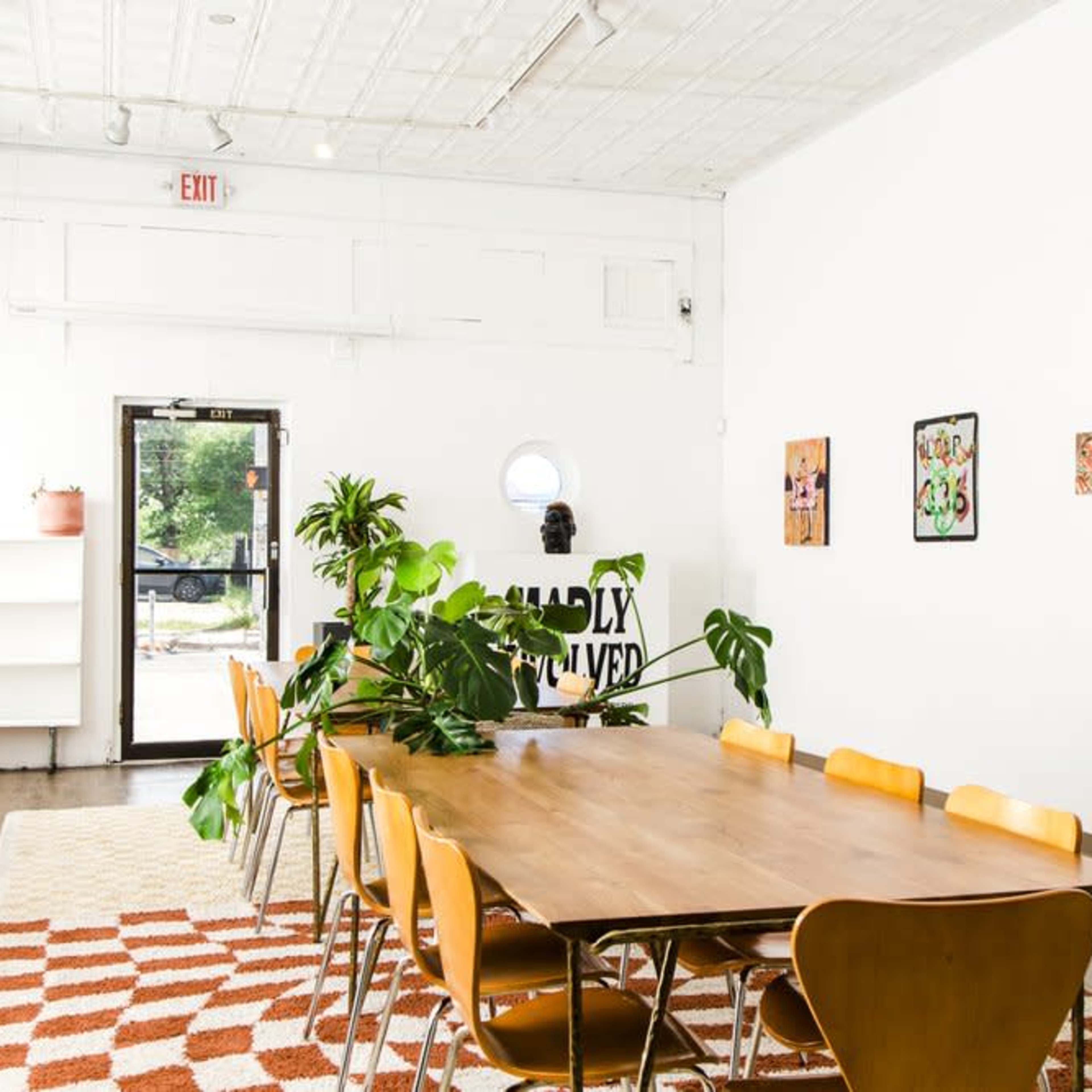 A spacious room features a wooden dining table surrounded by yellow chairs, adorned with plants and wall art, with a doorway leading outside.
