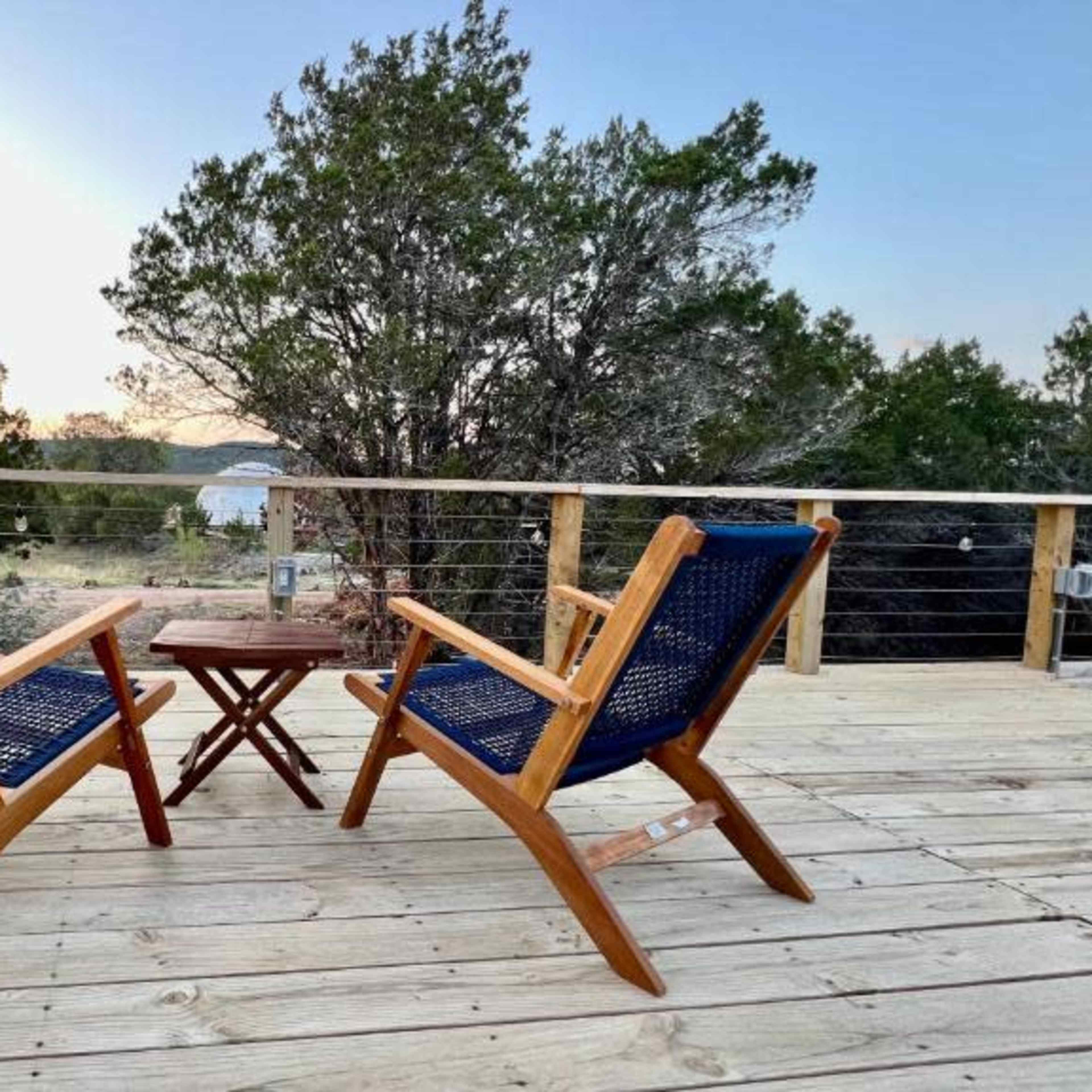 Two wooden chairs with blue woven seats are positioned around a small table on a wooden deck overlooking a scenic view of trees and water.