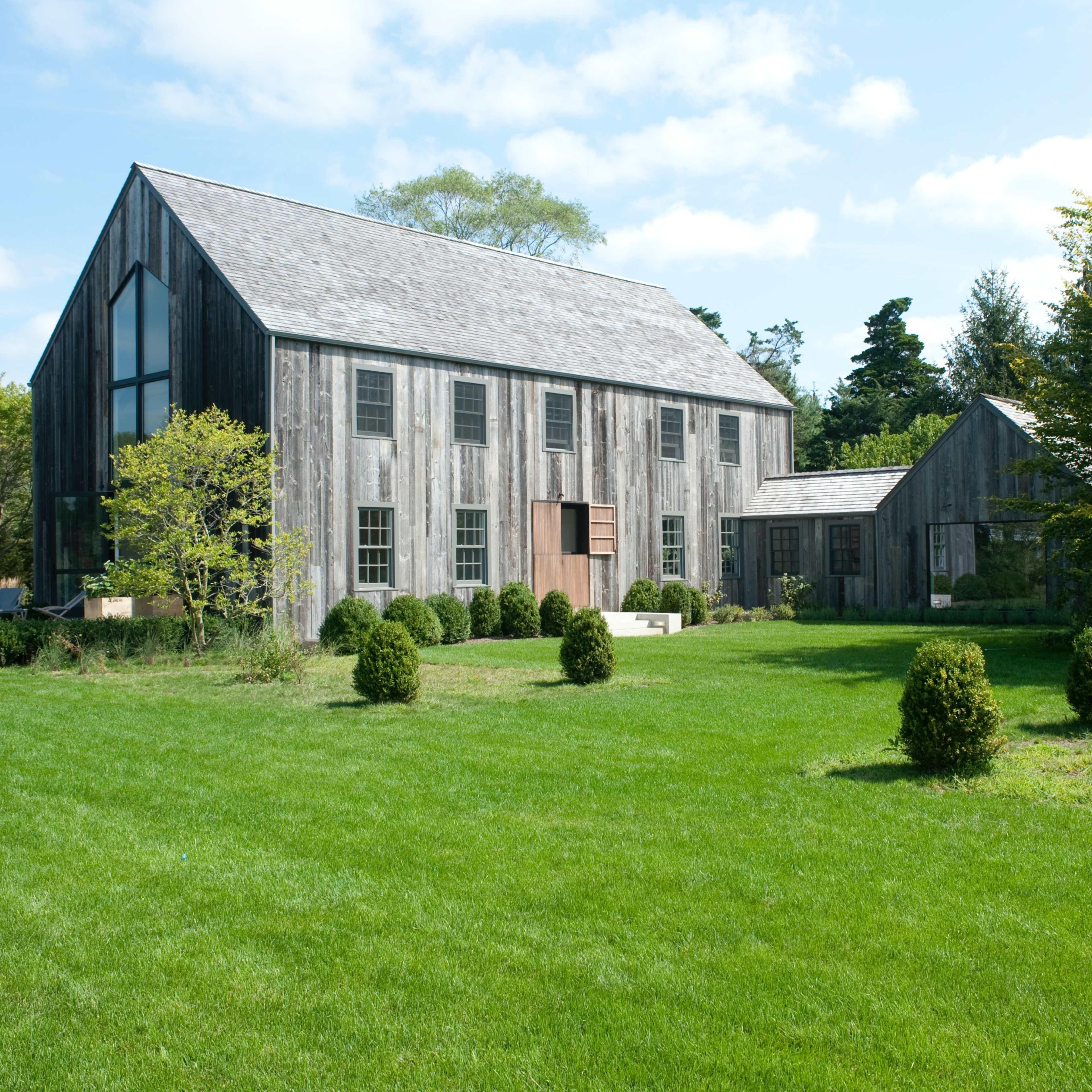 A large, contemporary barn-style building made of weathered wood is set on a green lawn surrounded by neatly trimmed bushes and trees.