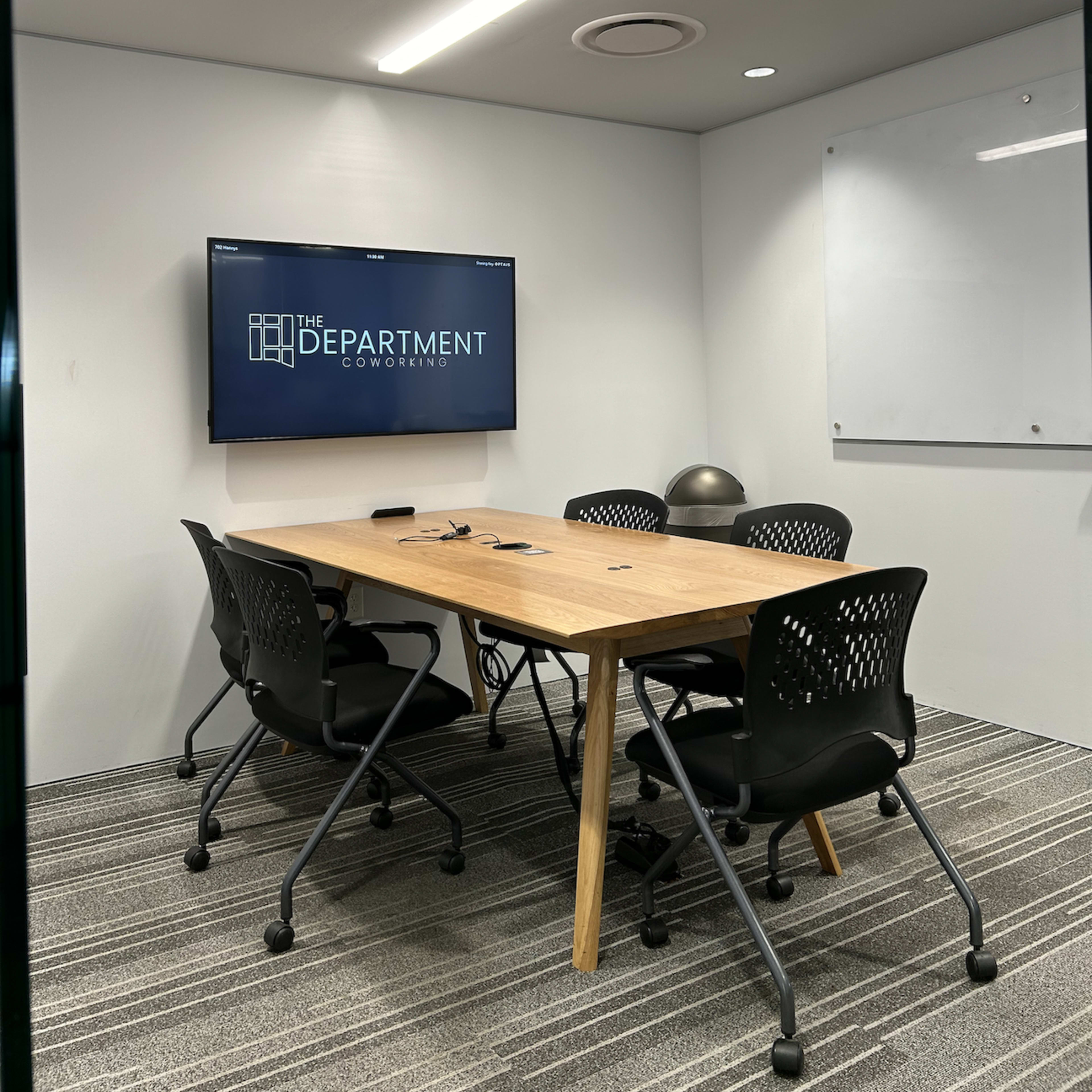 A modern conference room features a wooden table surrounded by four black office chairs, with a large screen displaying "THE DEPARTMENT" on the wall.