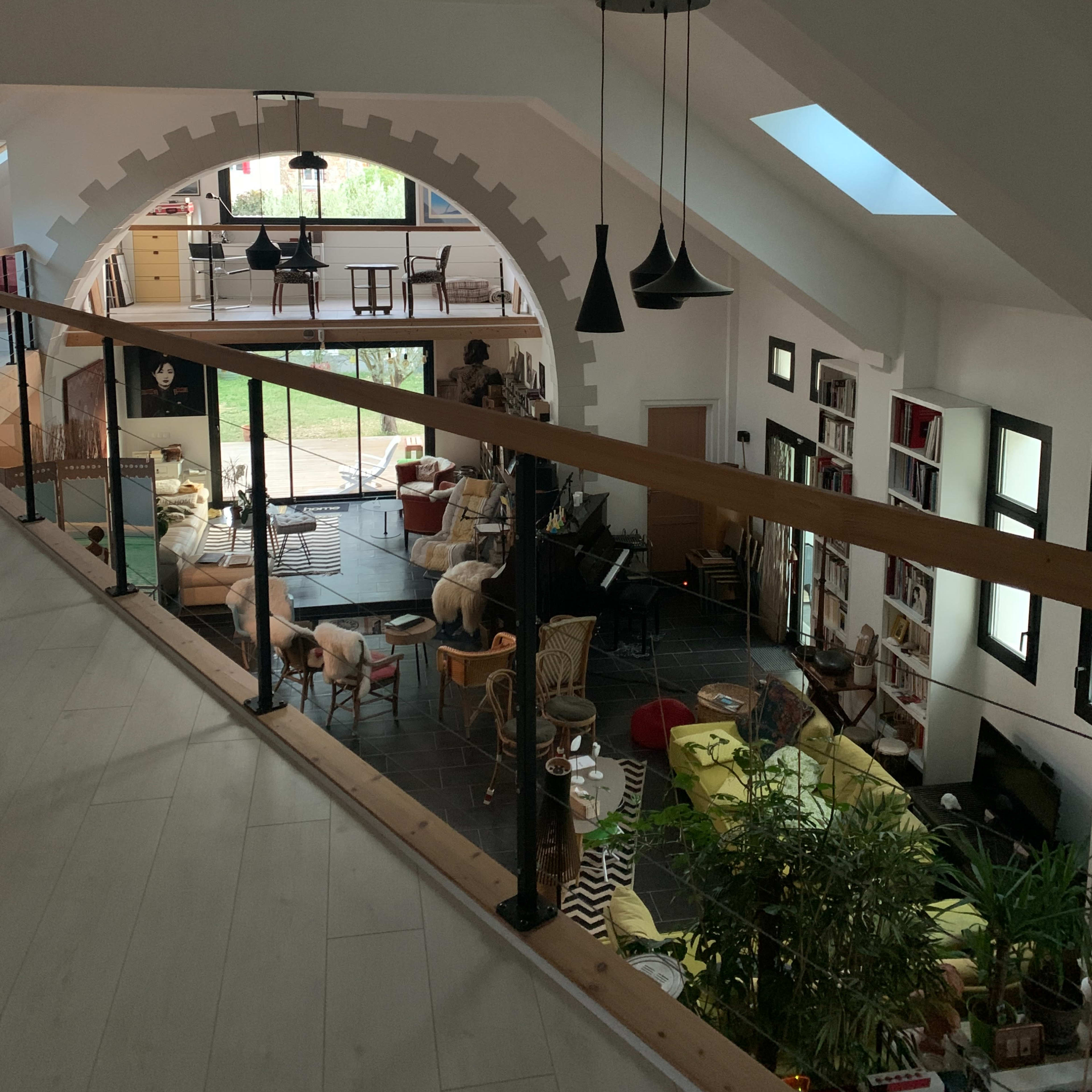 A spacious, modern interior features a balcony overlooking a lower living area filled with various furniture and plants, illuminated by natural light streaming through large windows.