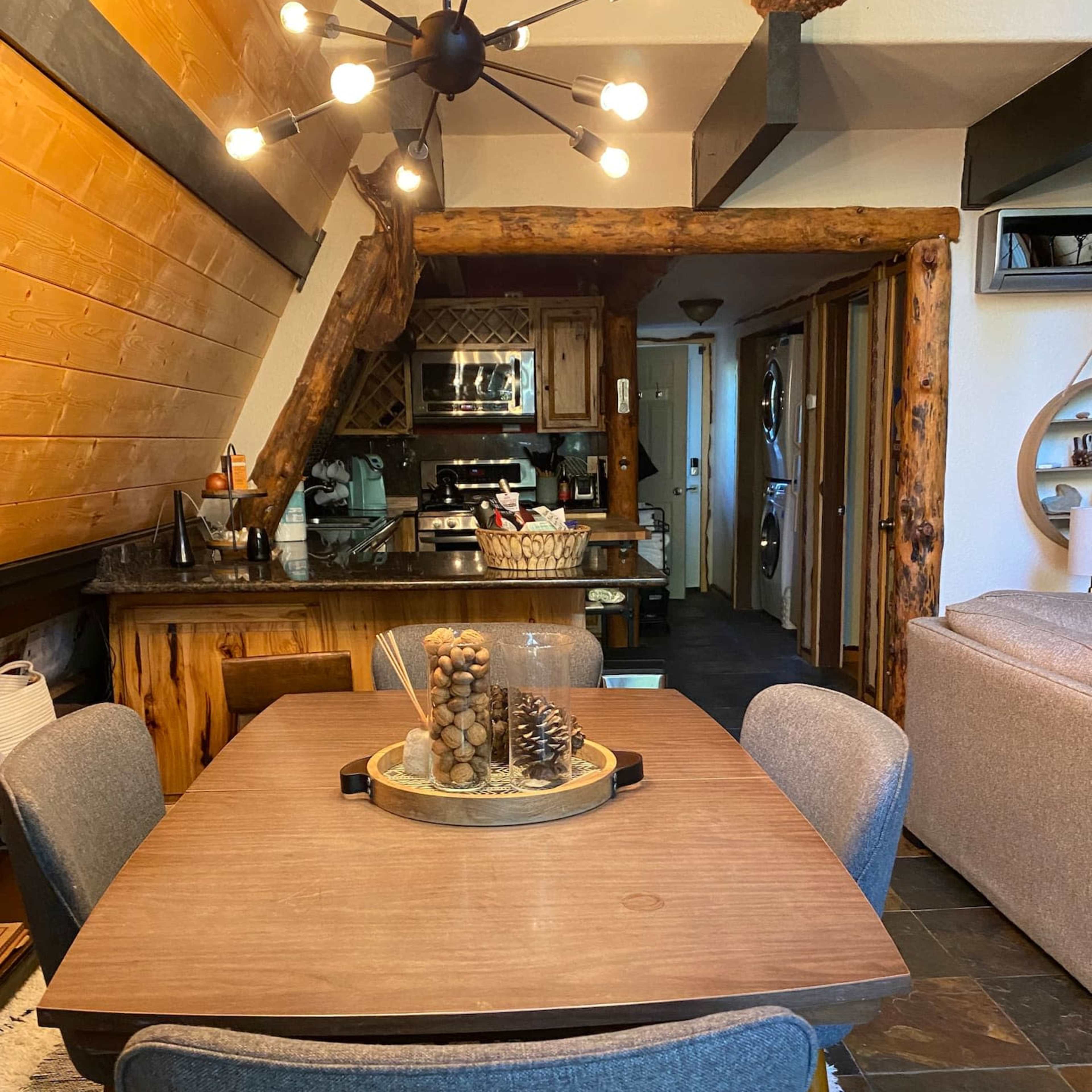 A dining area features a wooden table surrounded by upholstered chairs, with a central decorative tray and a backdrop of a kitchen visible through an archway.