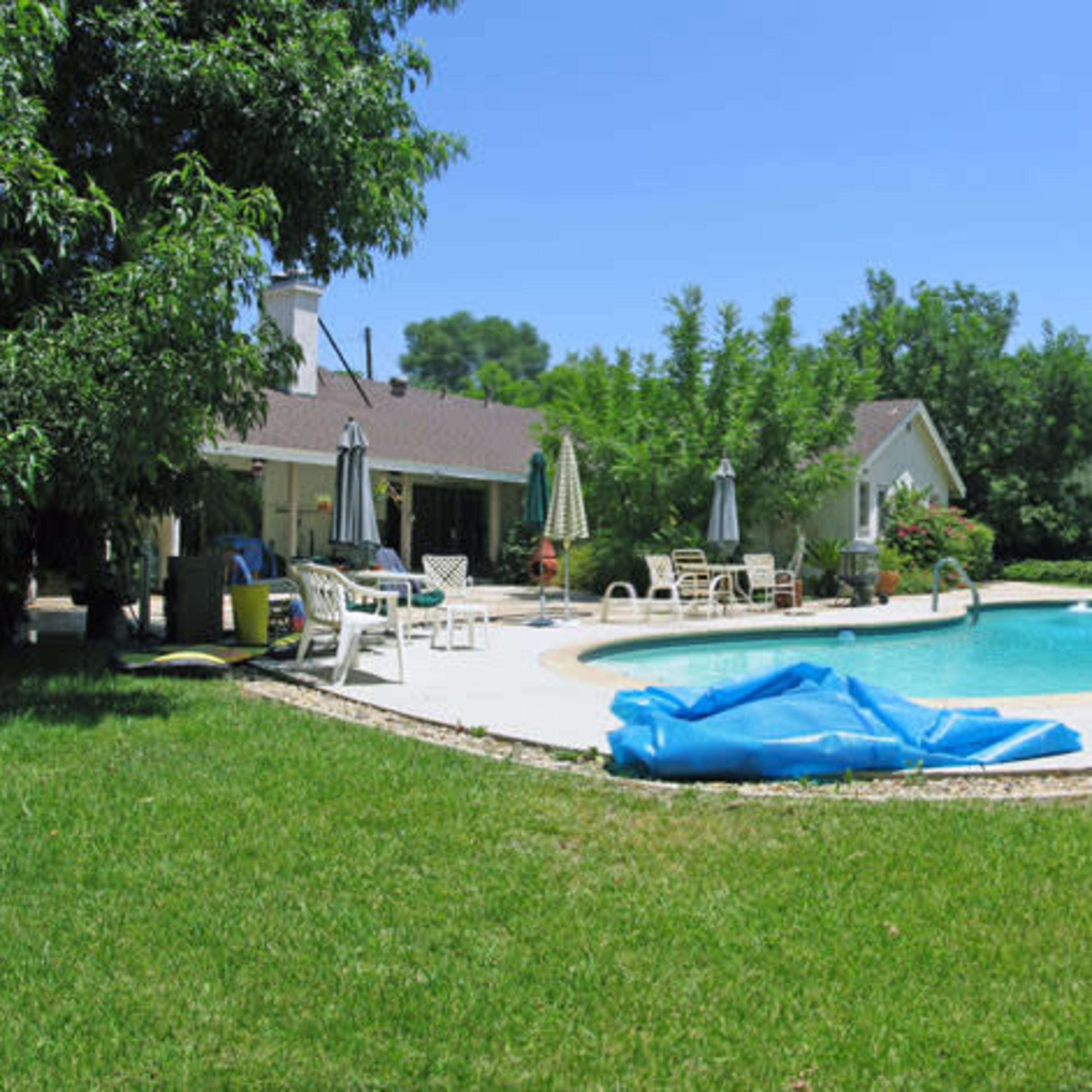 A backyard with a swimming pool surrounded by lounge chairs and shaded by trees.