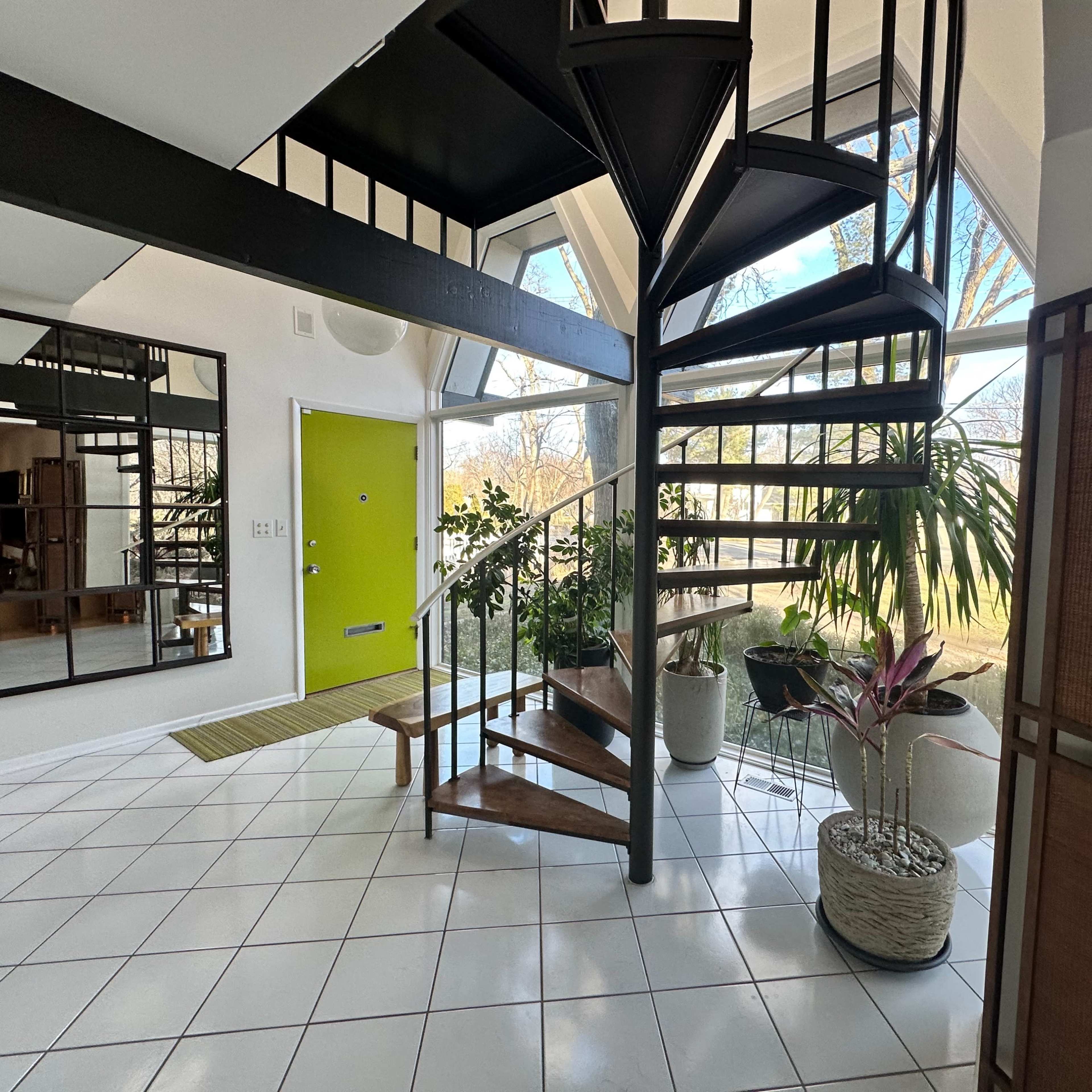 A modern entryway with a spiral staircase, large windows, and potted plants arranged around a tiled floor.