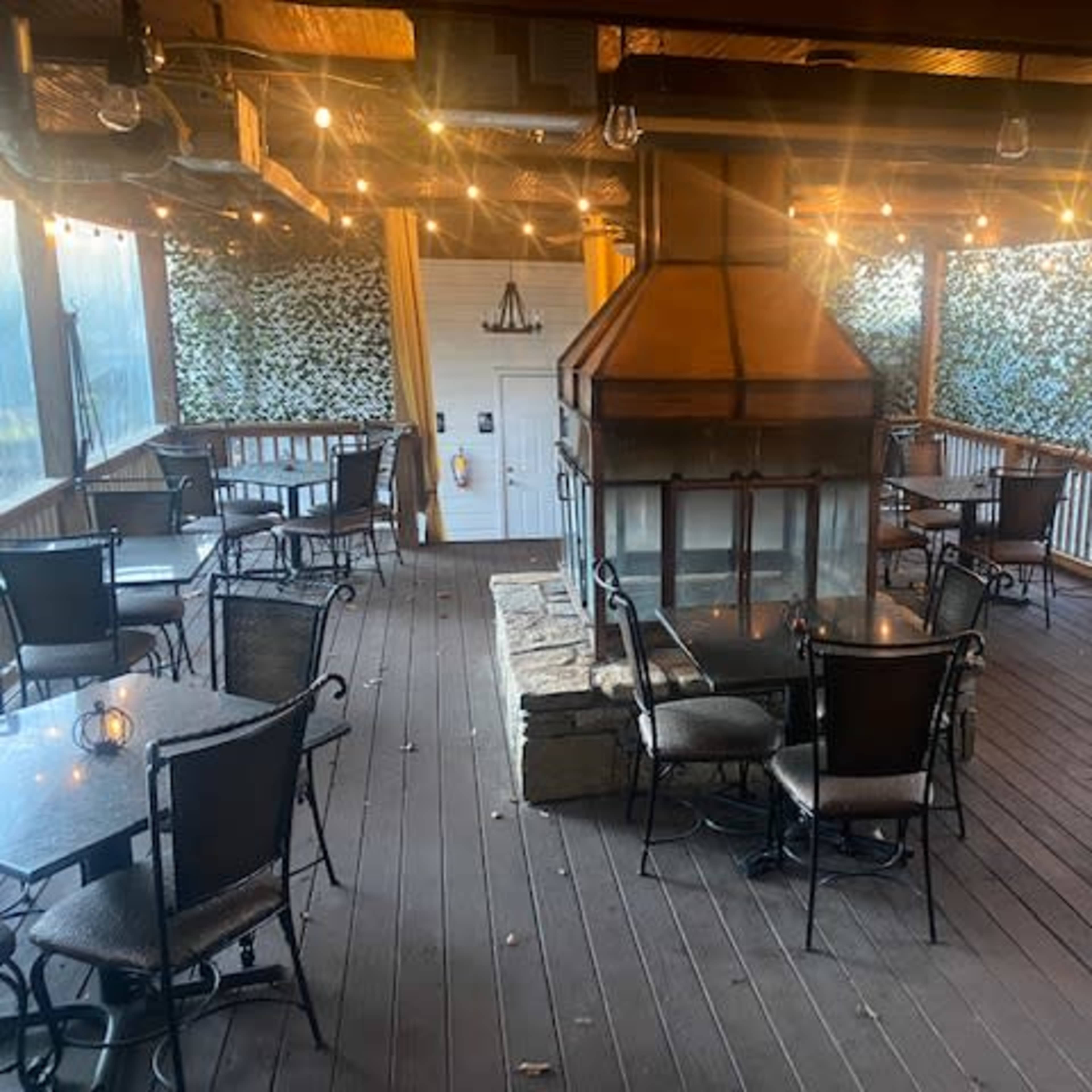 A spacious dining area with tables and chairs arranged around a central fireplace, surrounded by walls covered in greenery and string lights overhead.