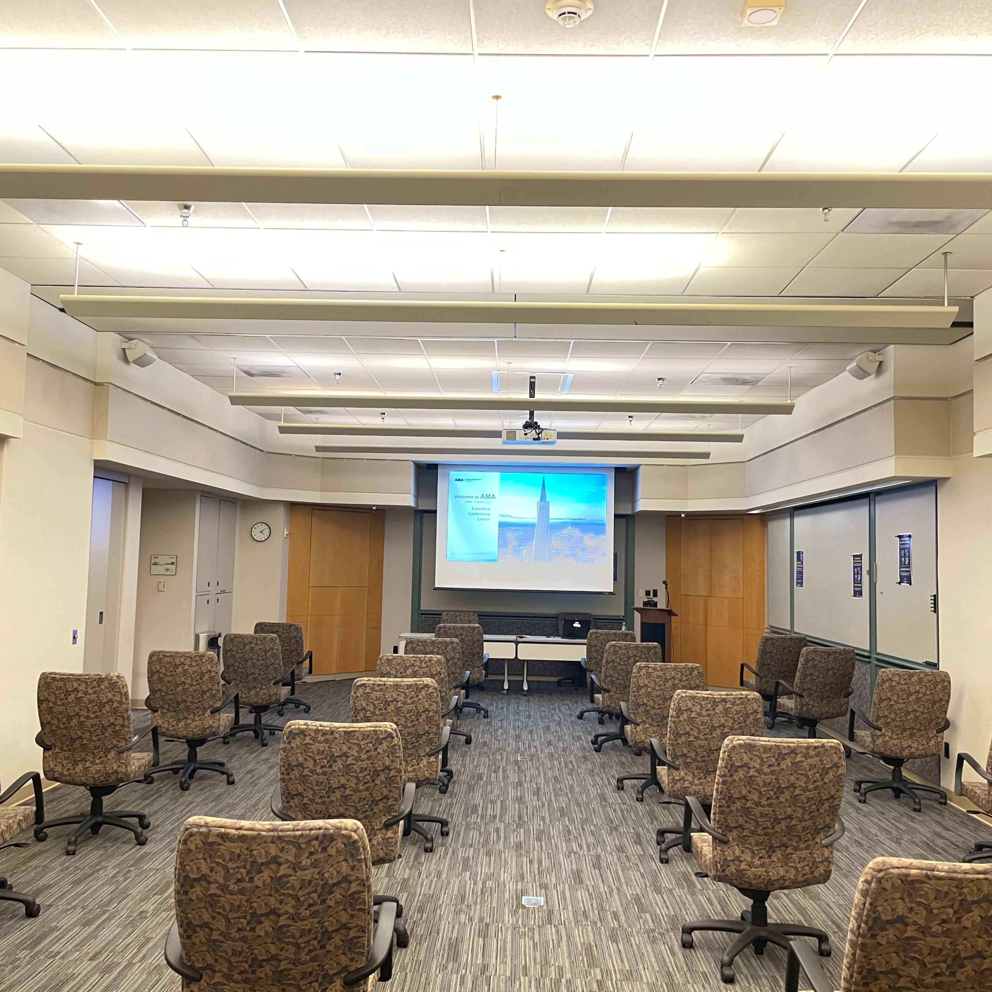 A conference room is set up with rows of rolling chairs facing a projector screen.