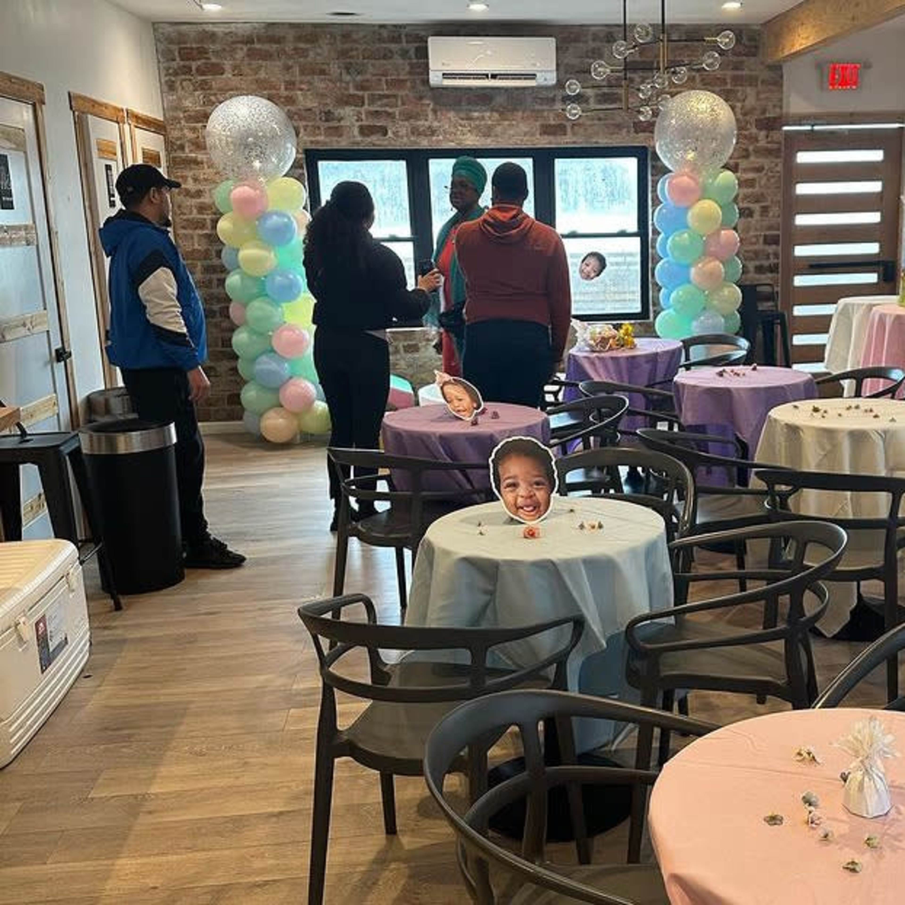 The interior of a decorated event space features tables covered with pastel-colored tablecloths, balloons, and playful cutouts of faces placed at each table.
