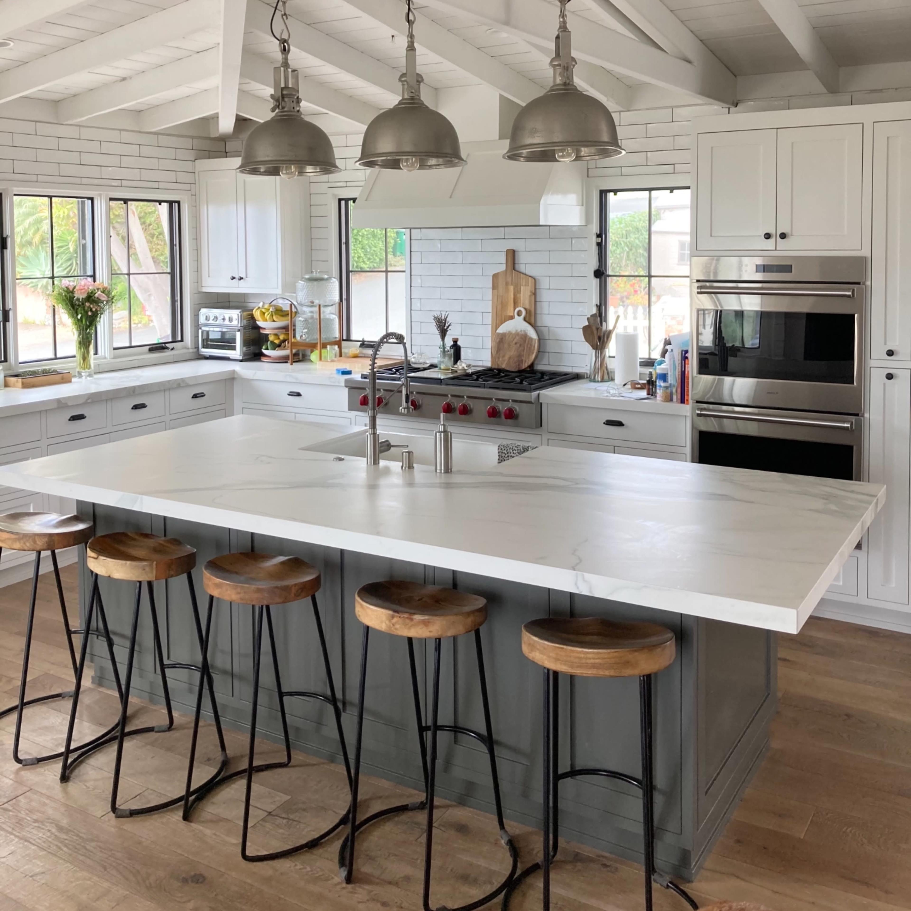 A modern kitchen with a large island, four wooden stools, and stainless steel appliances under white wooden beams.