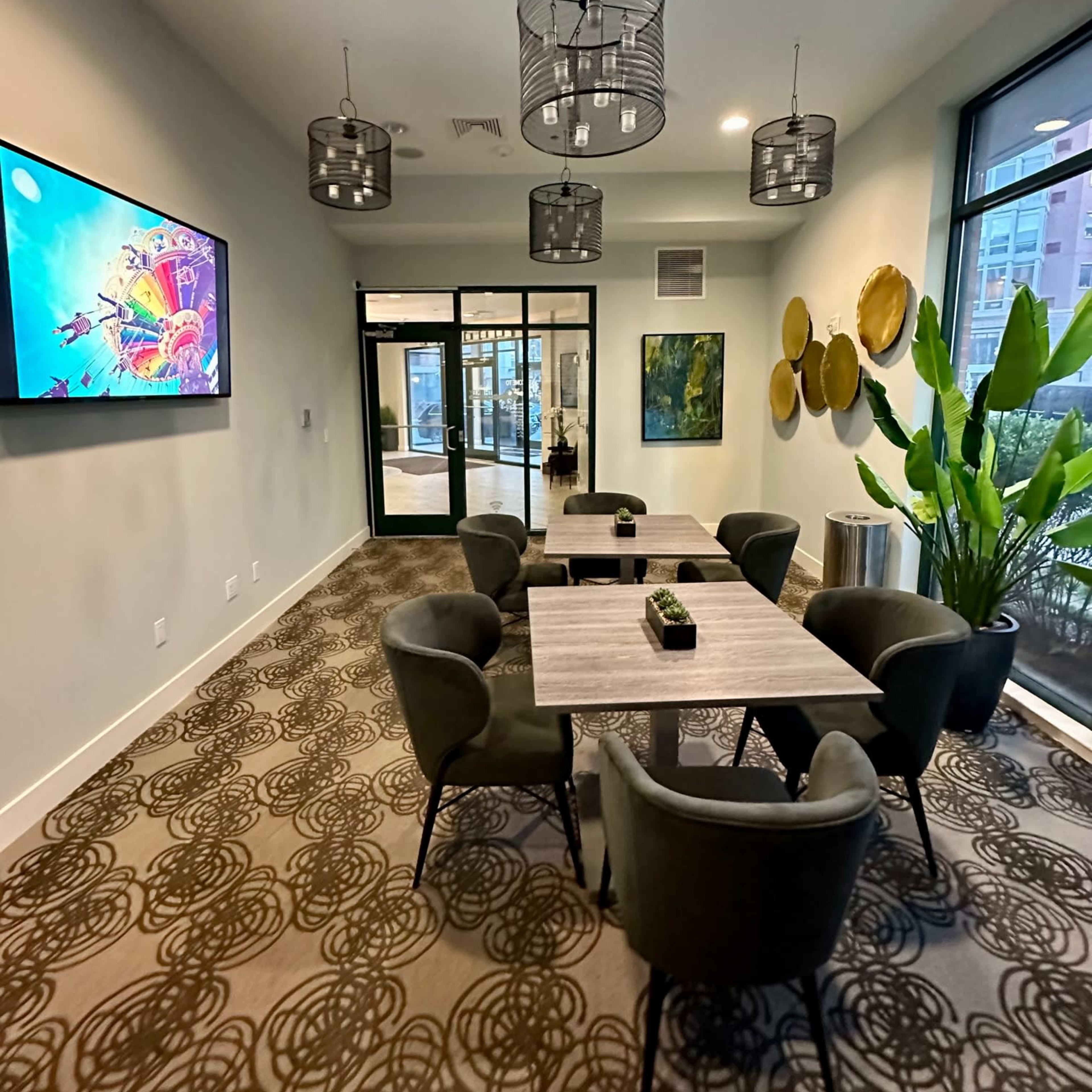 A well-lit meeting room features a large table surrounded by six chairs, with a television screen on the wall and greenery near the windows.