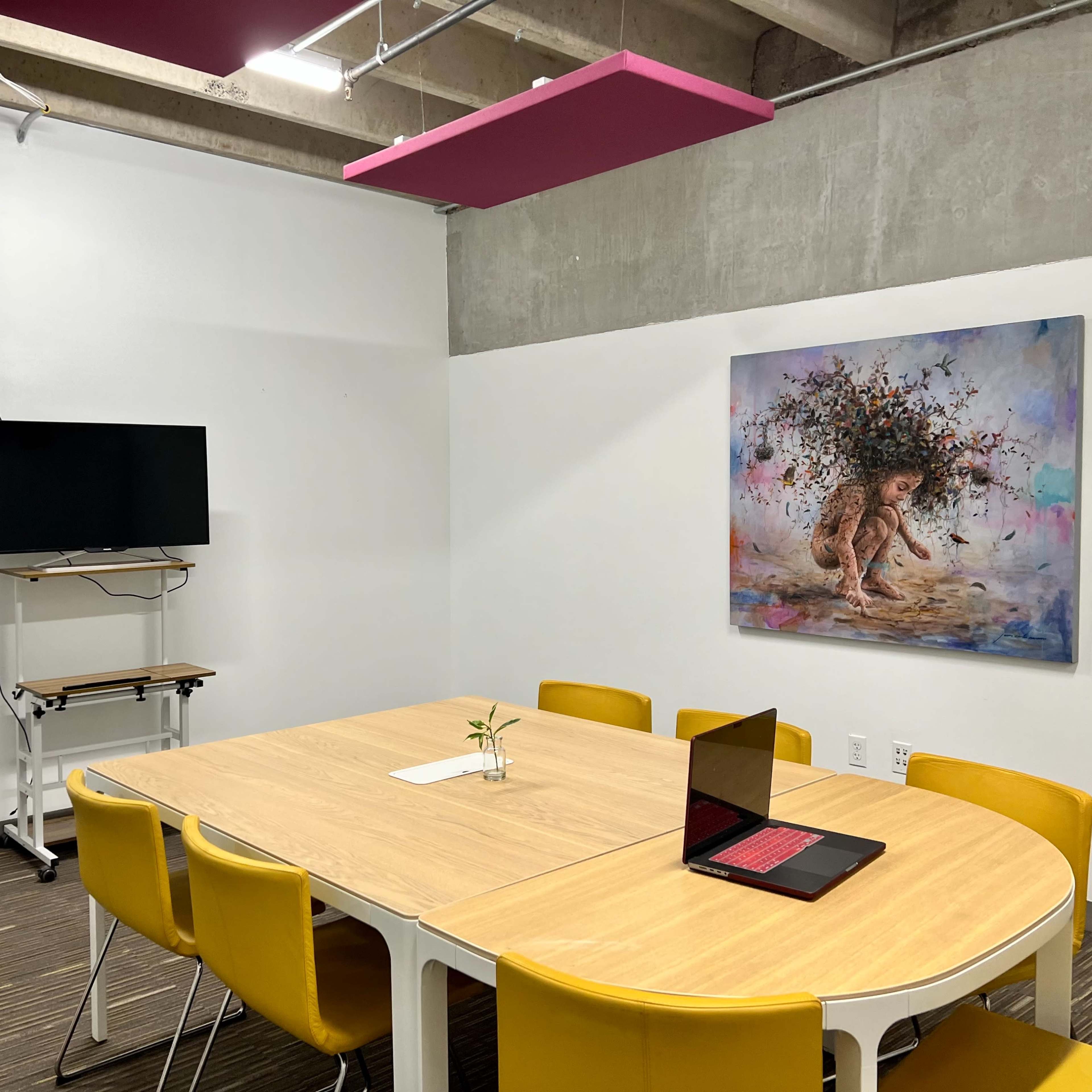 A meeting room features a wooden table surrounded by yellow chairs, a wall-mounted TV, and a colorful artwork depicting a lion.