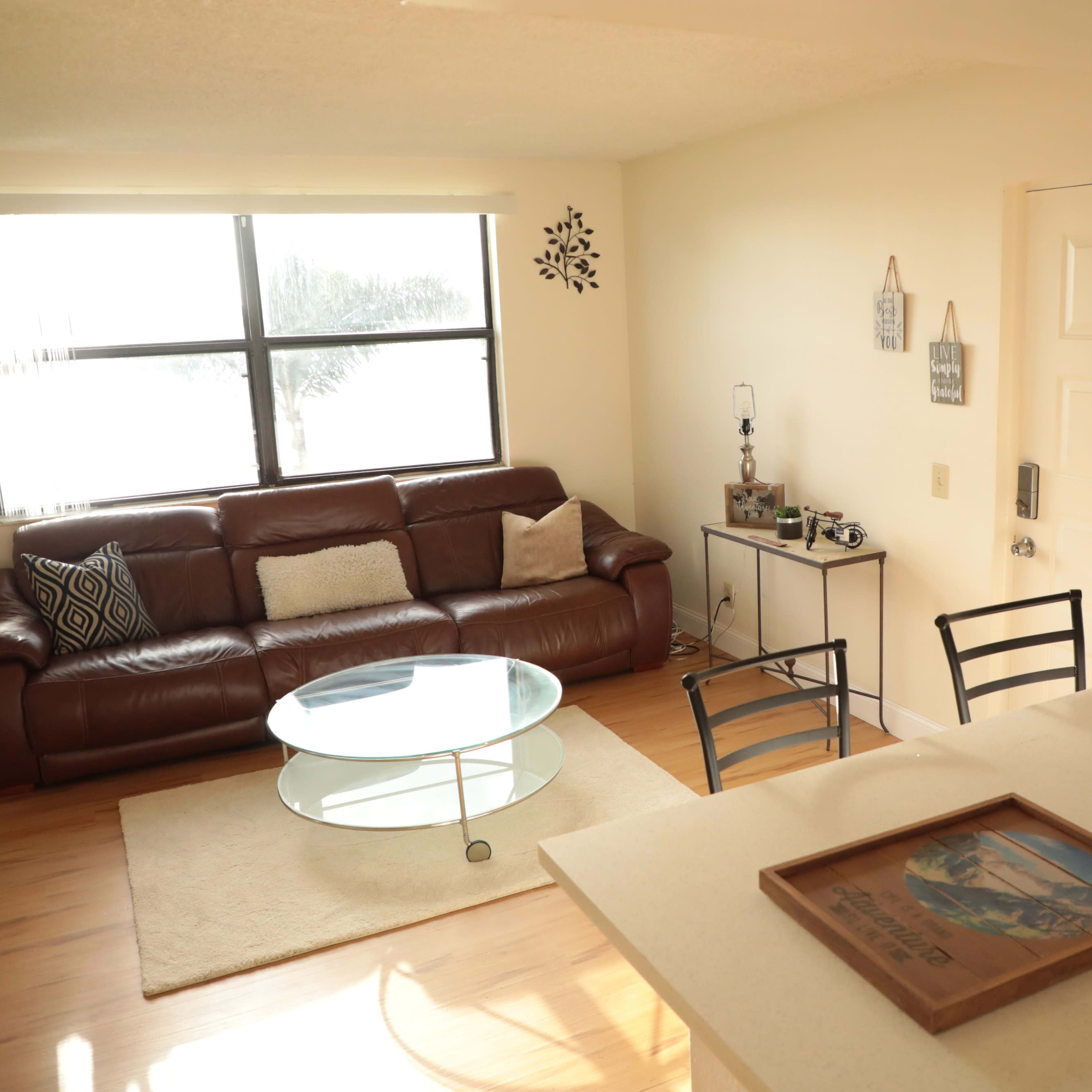 A living room with a brown leather sofa, a glass coffee table, and a small dining area with chairs, all illuminated by natural light from a window.