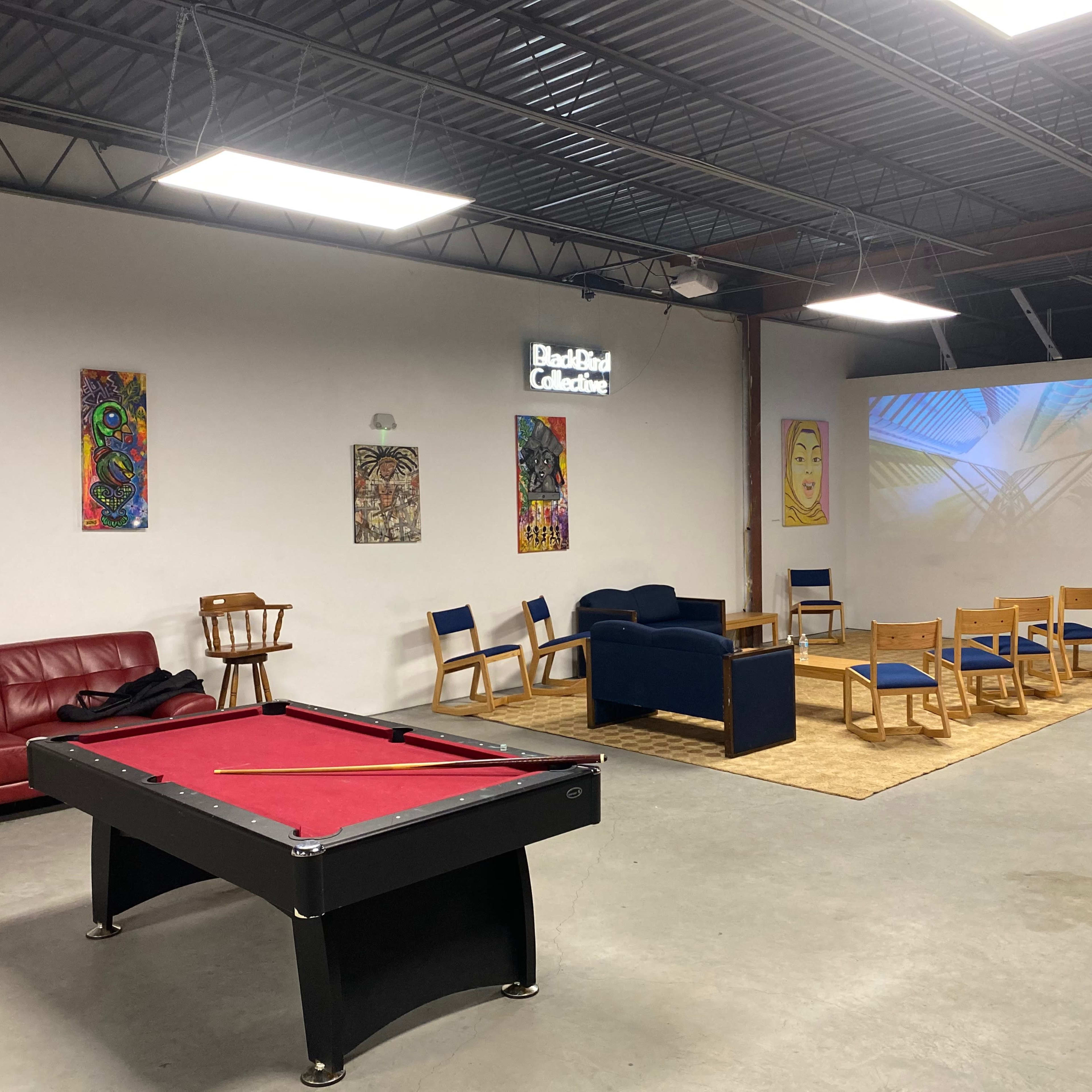 An indoor community space featuring a red pool table, a blue couch, several wooden chairs, and art displayed on the walls, with a projector screen showing a geometric design.