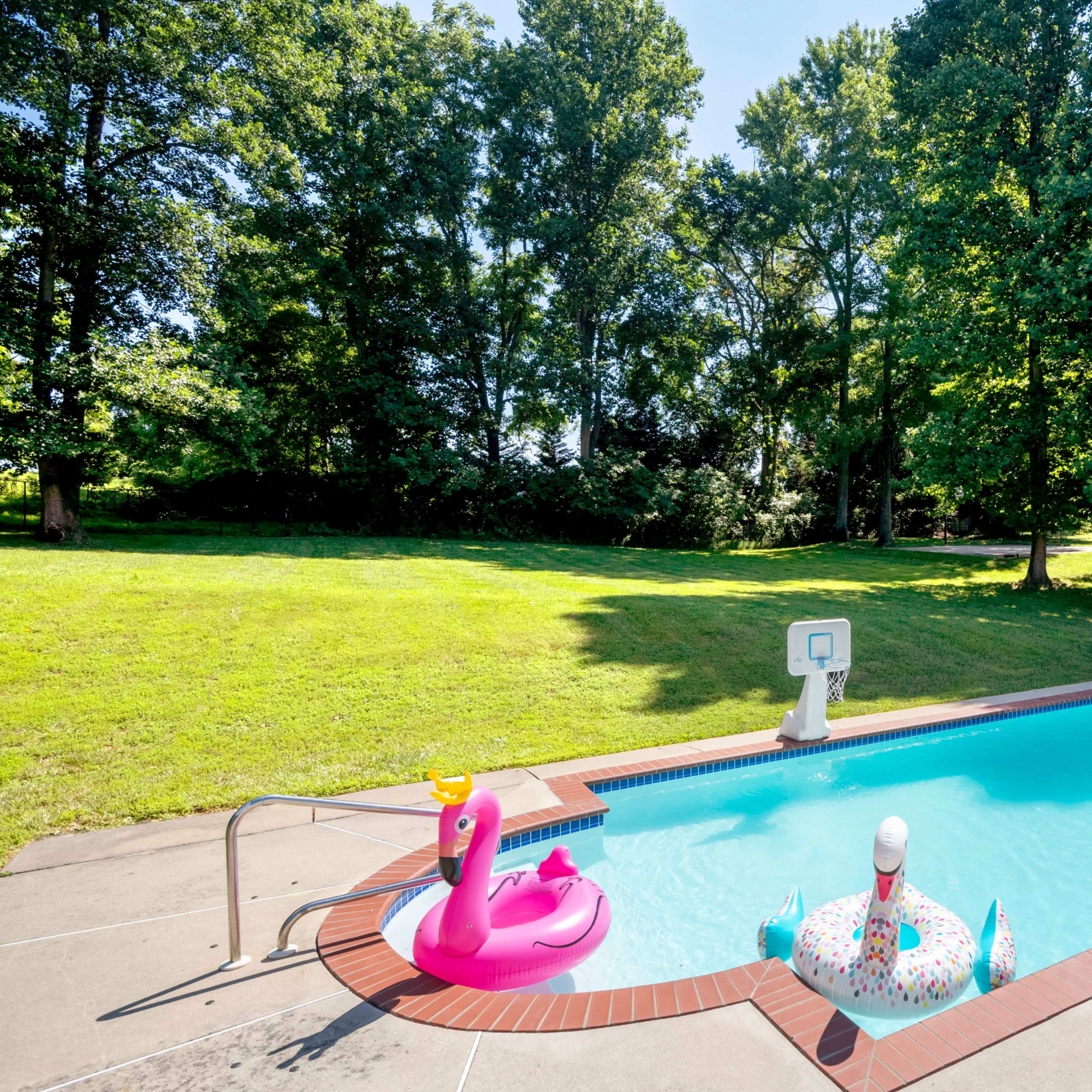 A swimming pool is surrounded by green grass and trees, featuring inflatable floats shaped like a flamingo and a unicorn.
