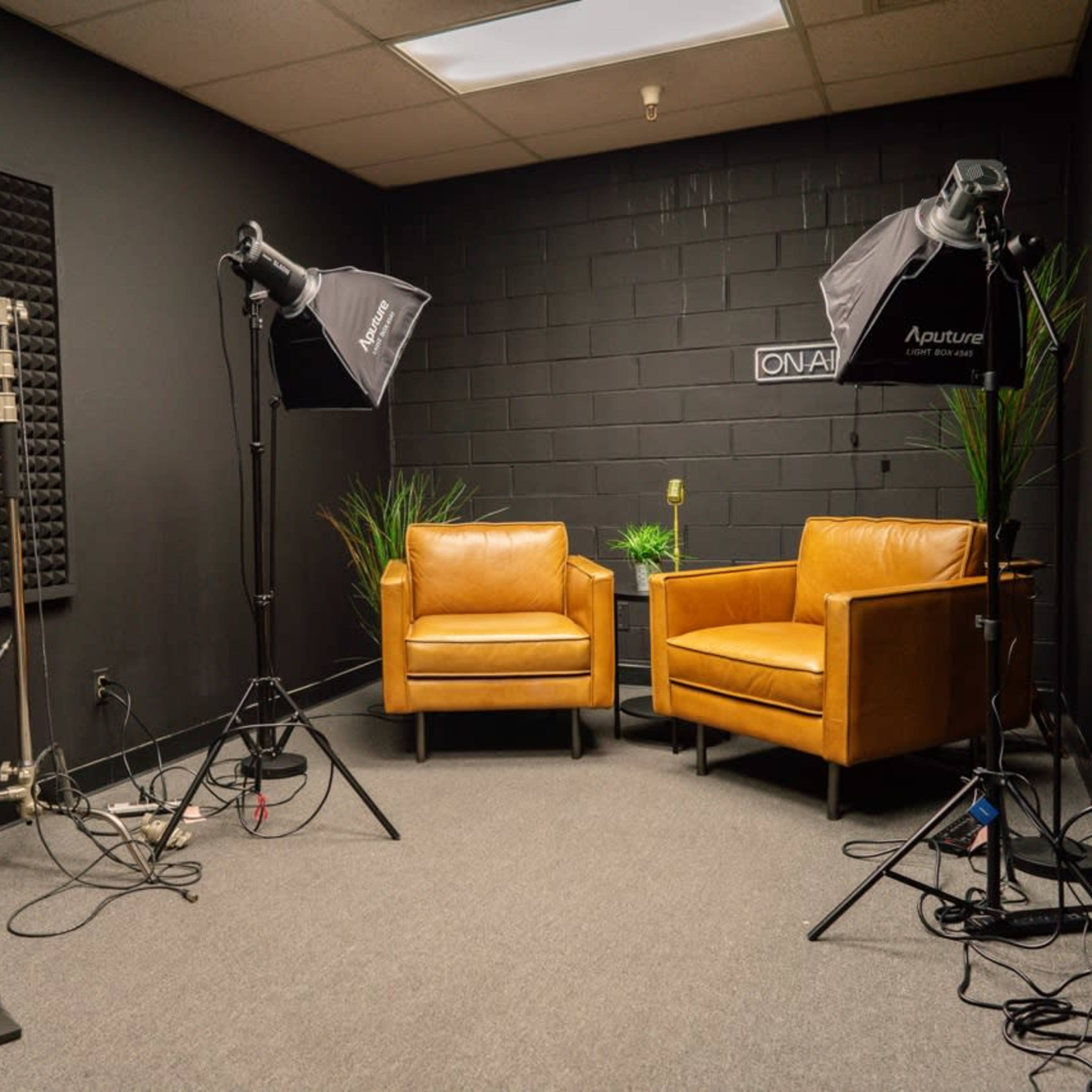 A small, dimly lit studio with two brown chairs, lighting equipment, and soundproof foam panels on the walls.