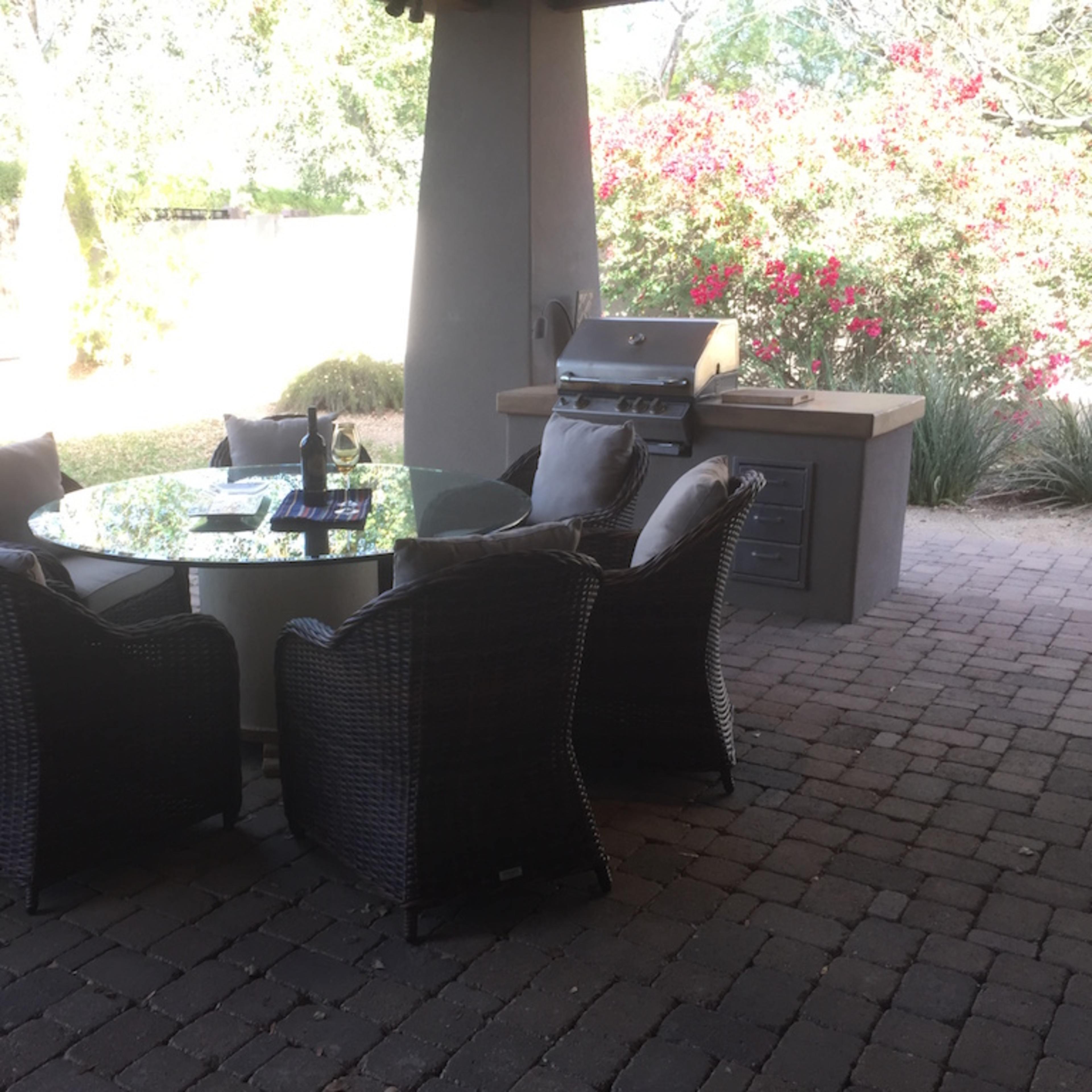 A patio area features a circular table surrounded by four wicker chairs, with a barbecue grill and flowering bushes in the background.