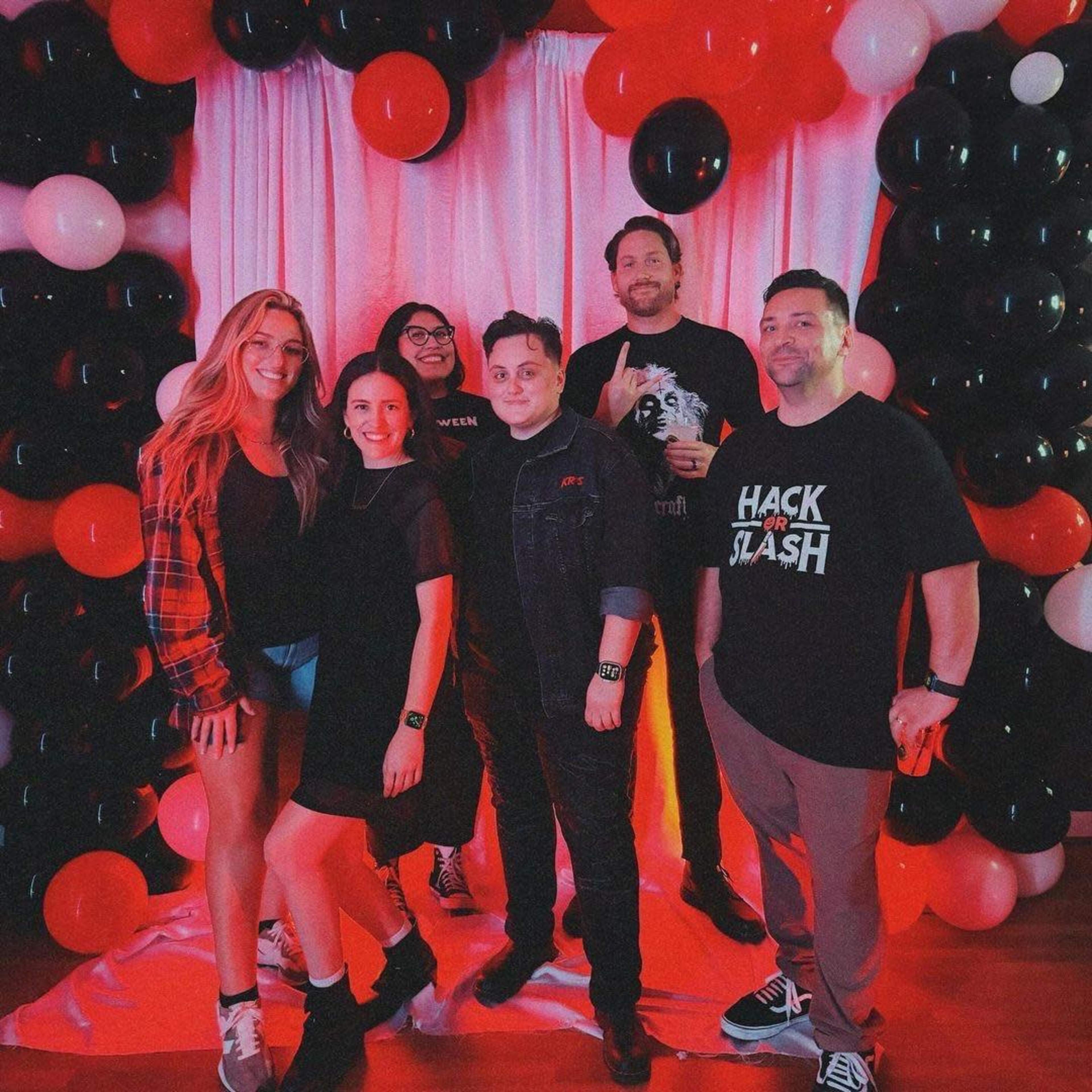 A group of six people poses in front of a balloon arch made of black, red, and white balloons.