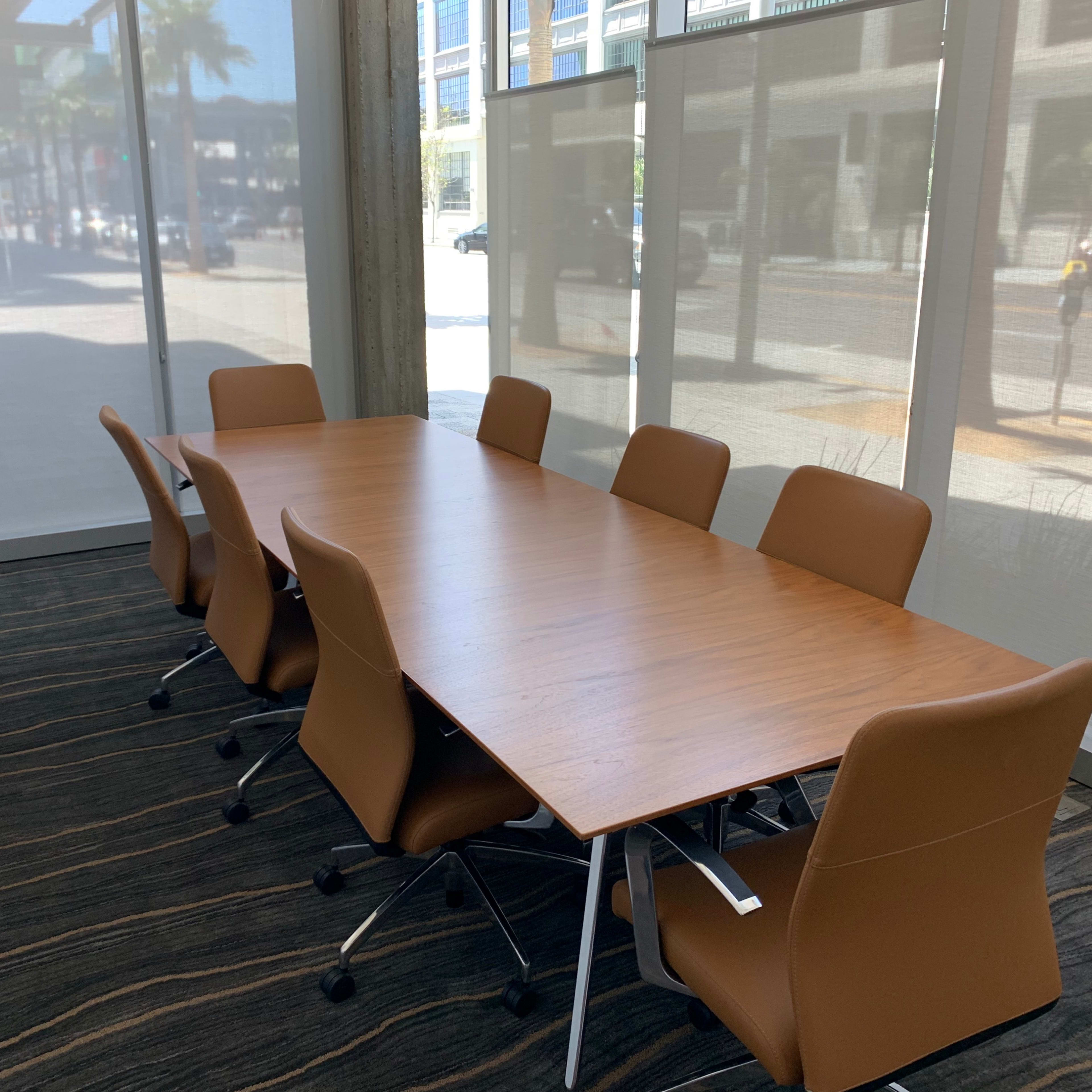 A long wooden conference table is set with brown chairs in a well-lit meeting room with large windows.