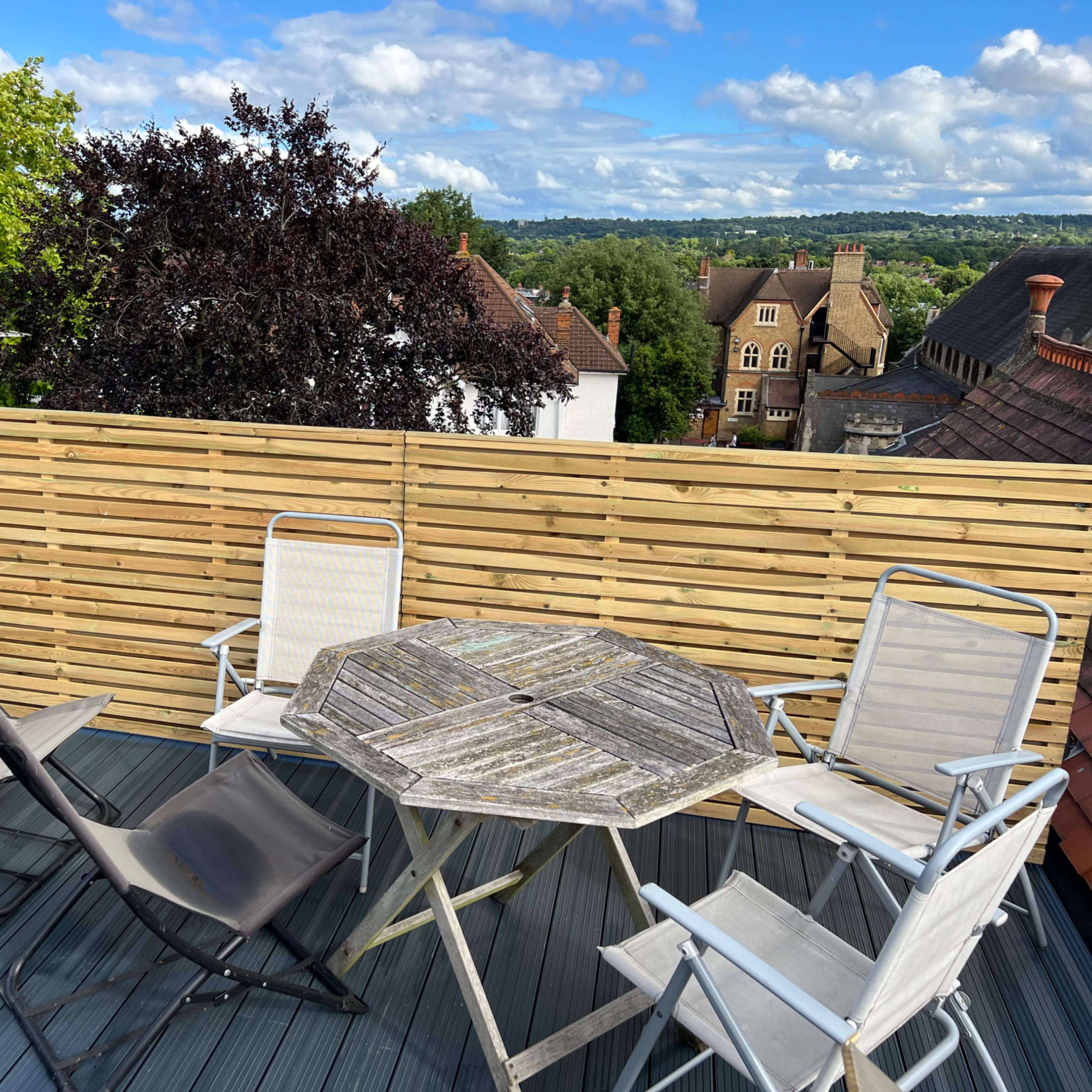 A wooden patio table surrounded by four chairs is set on a deck, overlooking rooftops and trees under a partly cloudy sky.