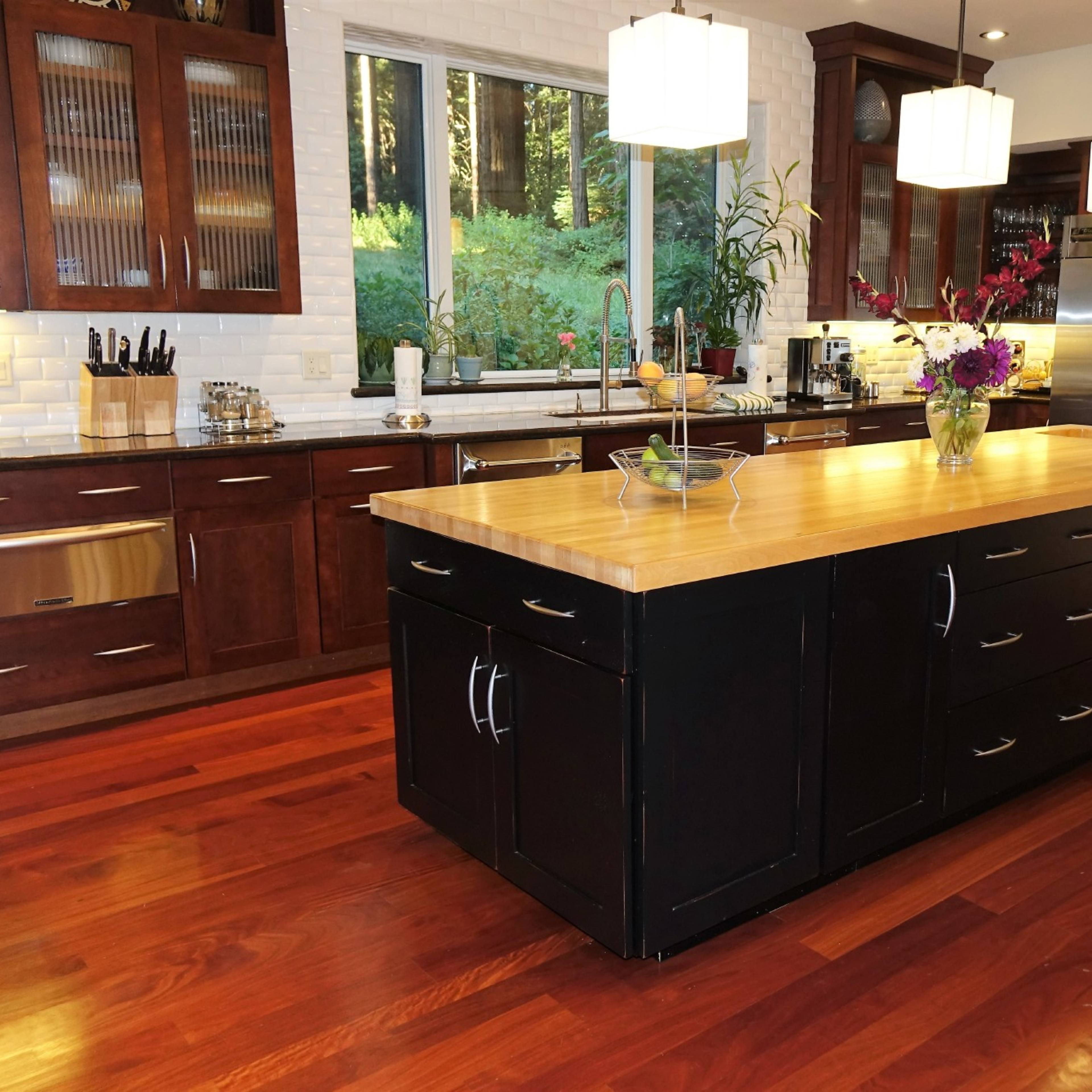 A modern kitchen features a large wooden island with drawers, surrounded by dark cabinetry and stainless steel appliances, complemented by large windows that provide views of a forest.