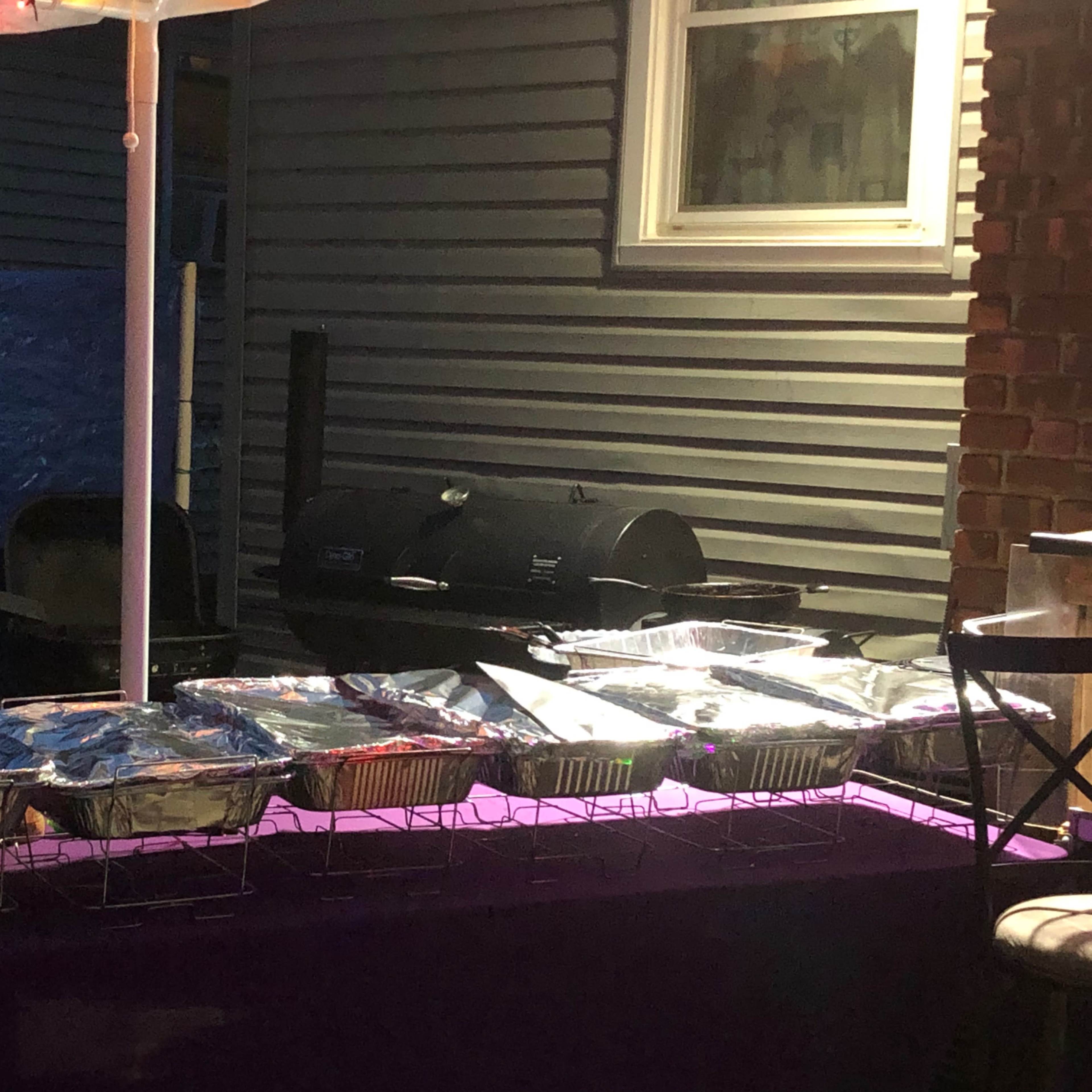 A row of covered trays is set on a table next to a grill and a blue tarp in a backyard setting at night.