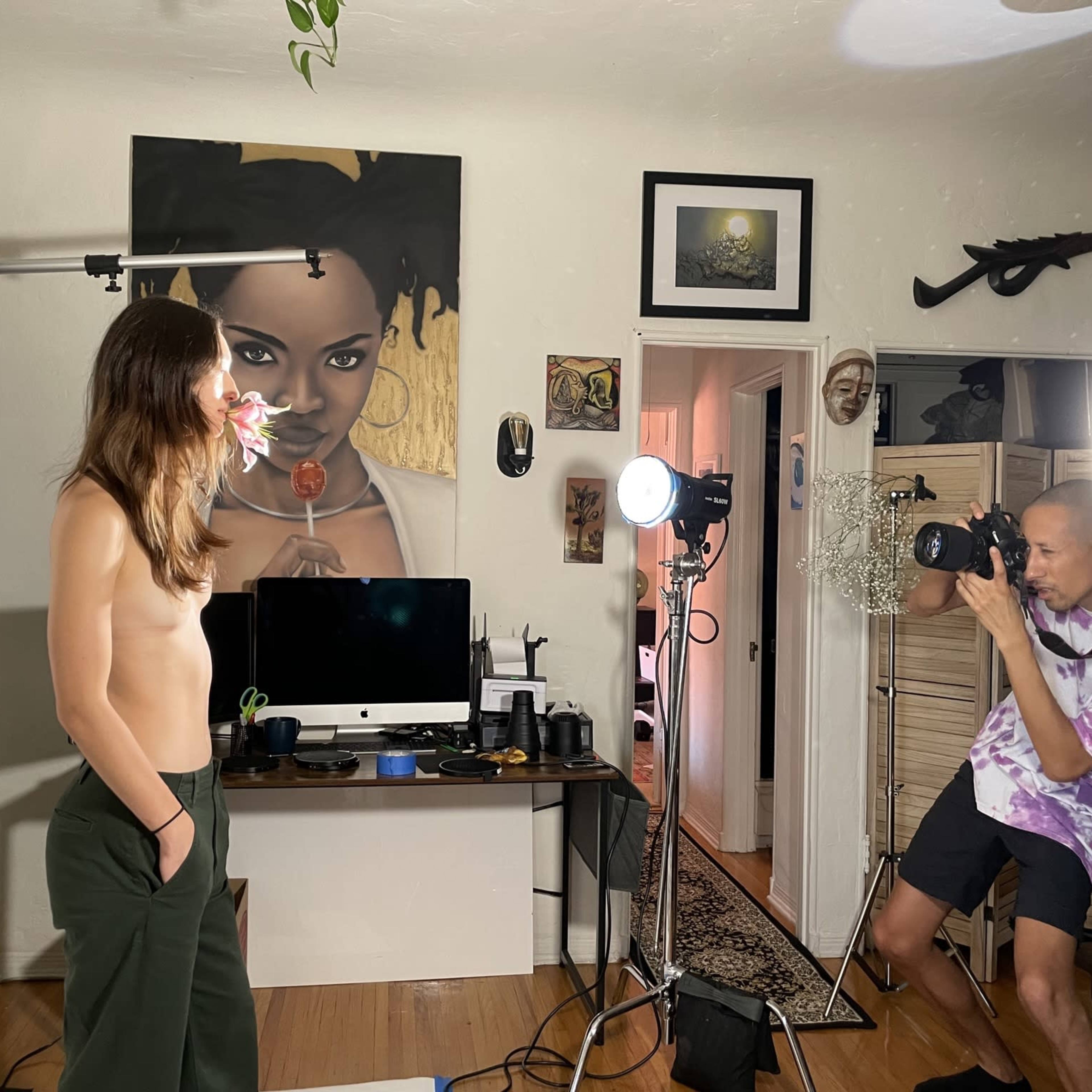 A shirtless model stands in front of a backdrop while a photographer captures the scene, with a large painting of a woman on the wall behind them.