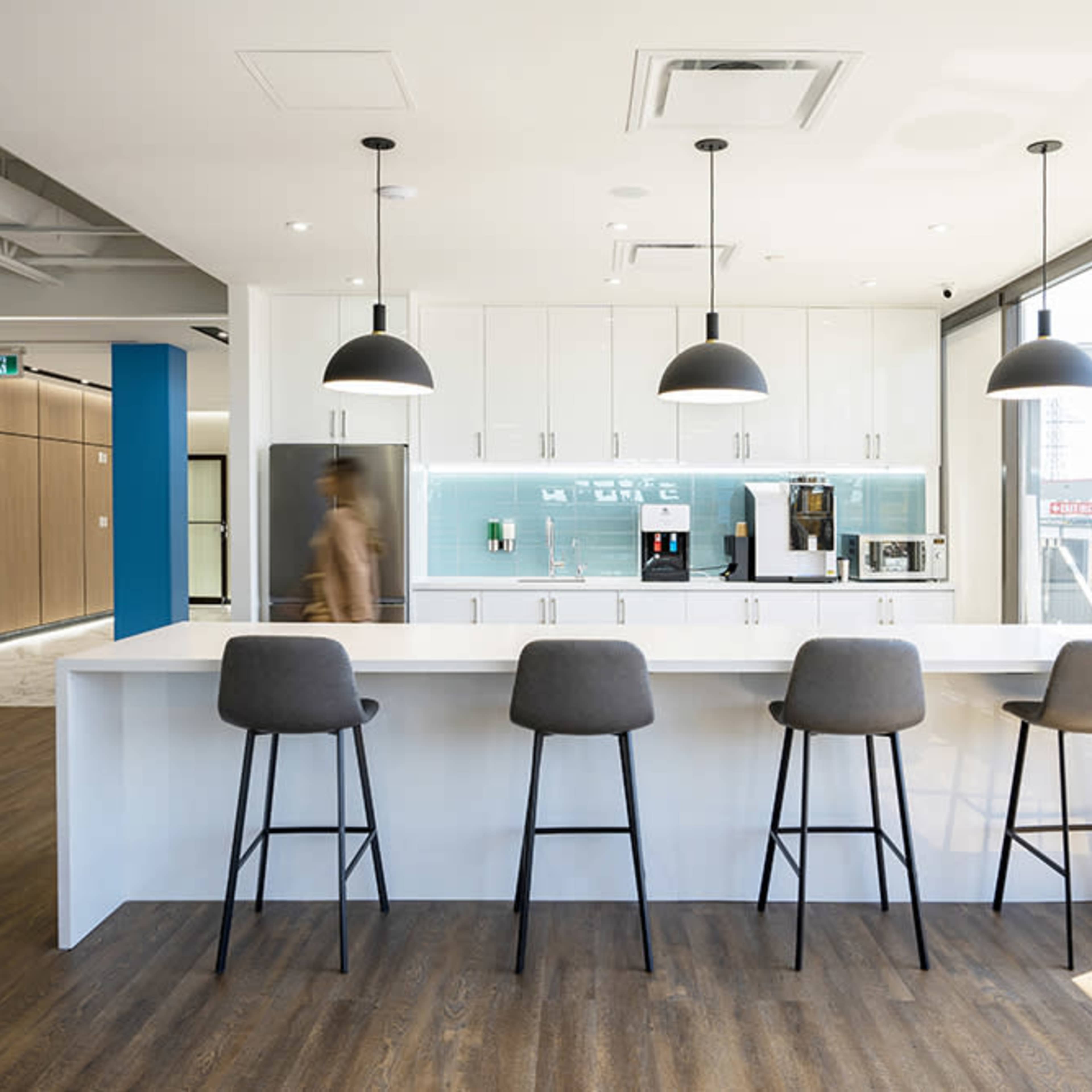 A modern kitchen with a white countertop and four bar stools, featuring a coffee machine and other appliances against a light blue backsplash.