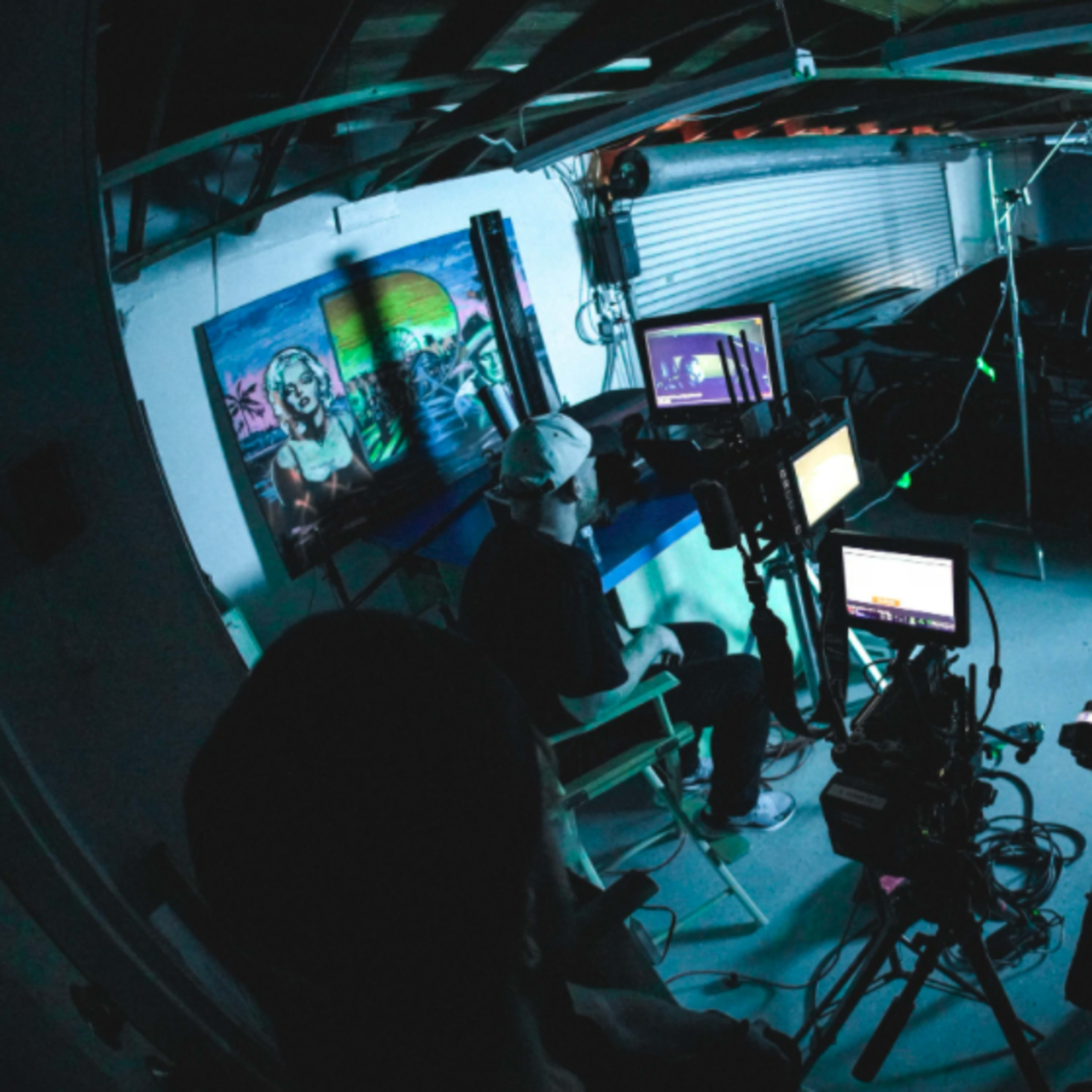 A person sits in a chair next to a black car in a dimly lit garage with multiple cameras and screens set up for filming.