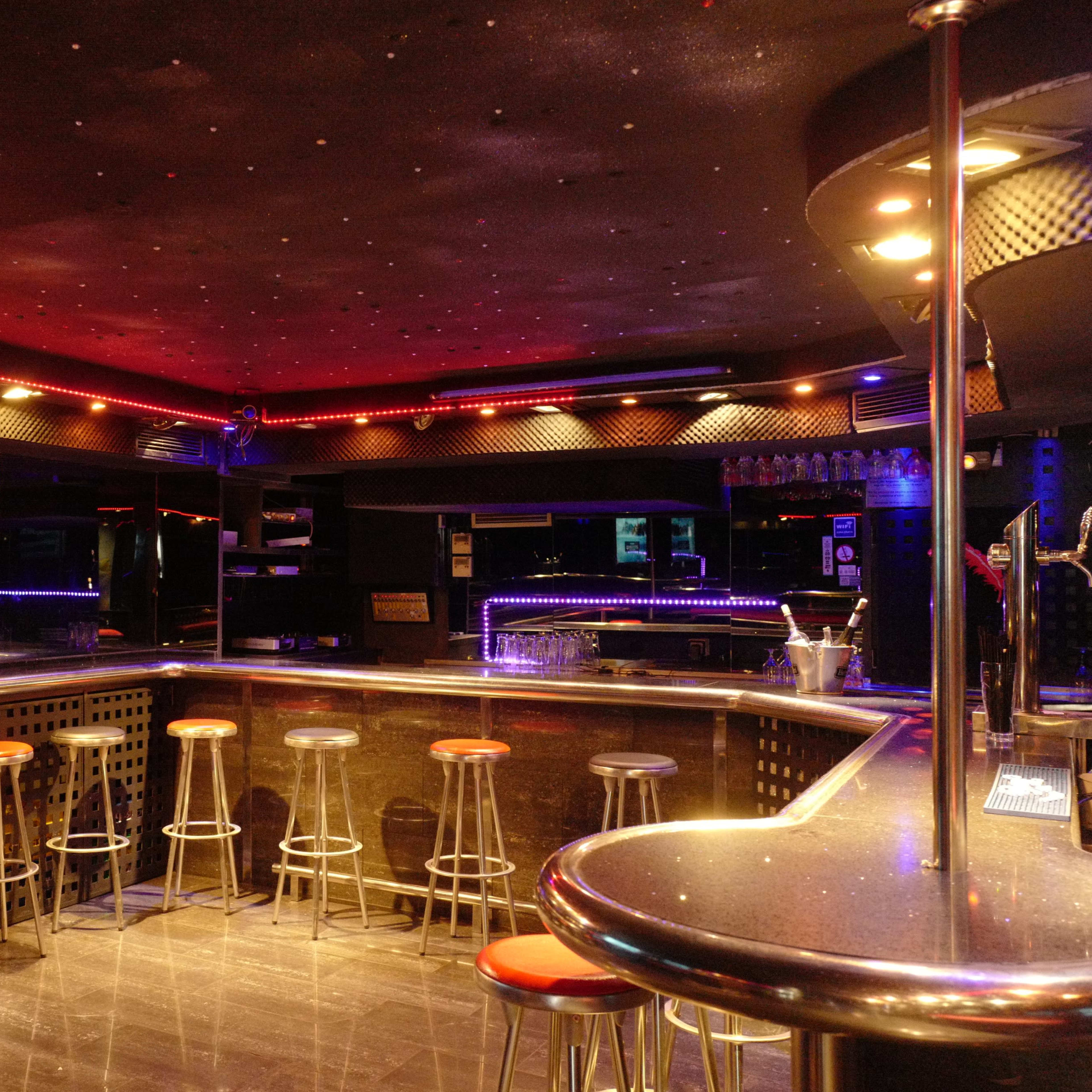 An empty bar area with high stools and a lit counter, featuring colorful lighting along the walls and ceiling.