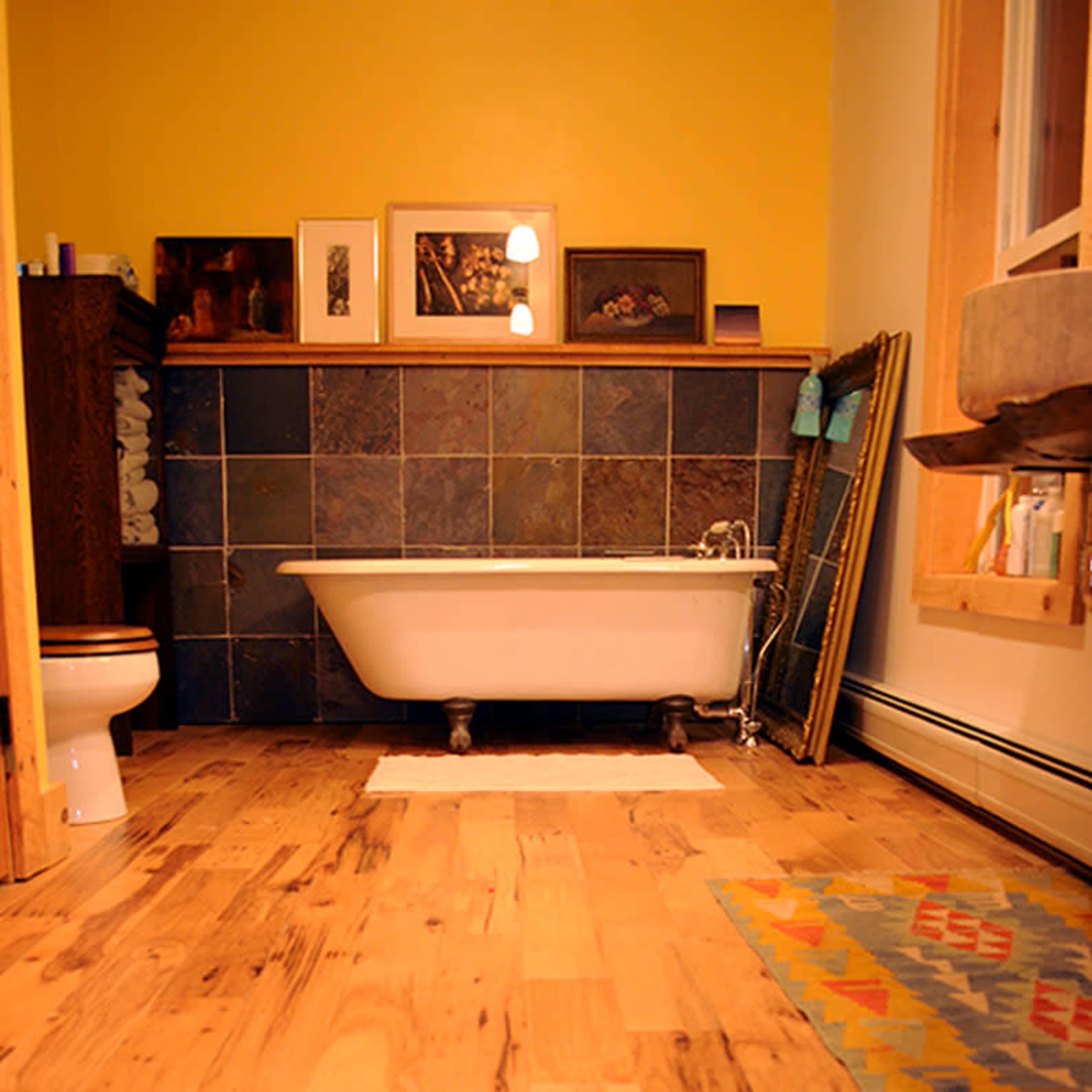 A bathroom with a wooden floor, a white bathtub, a toilet, and colorful wall art above a tiled backsplash.
