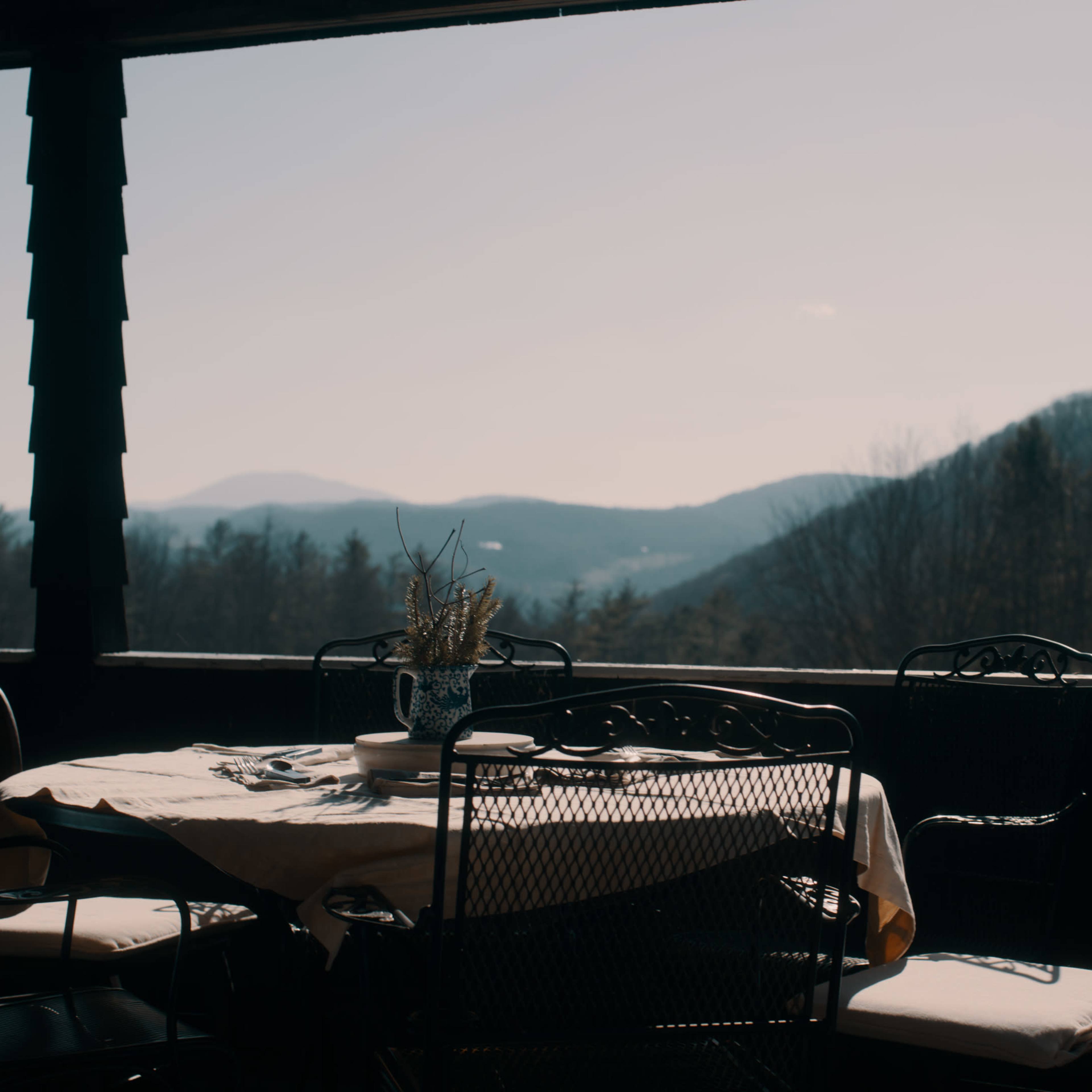 A table set with chairs overlooks a mountainous landscape from a porch.