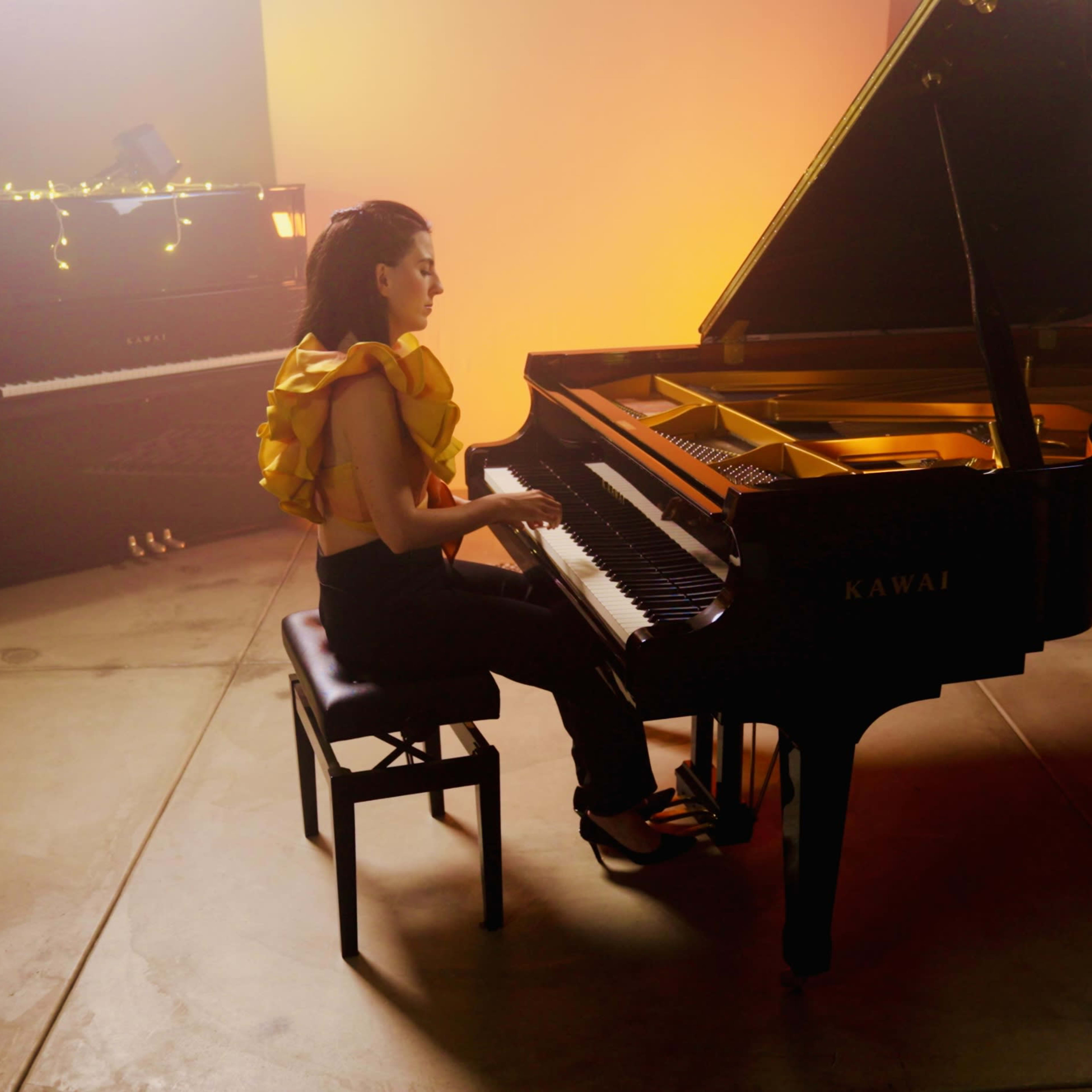 A woman in a ruffled yellow top plays a grand piano in a warmly lit room.