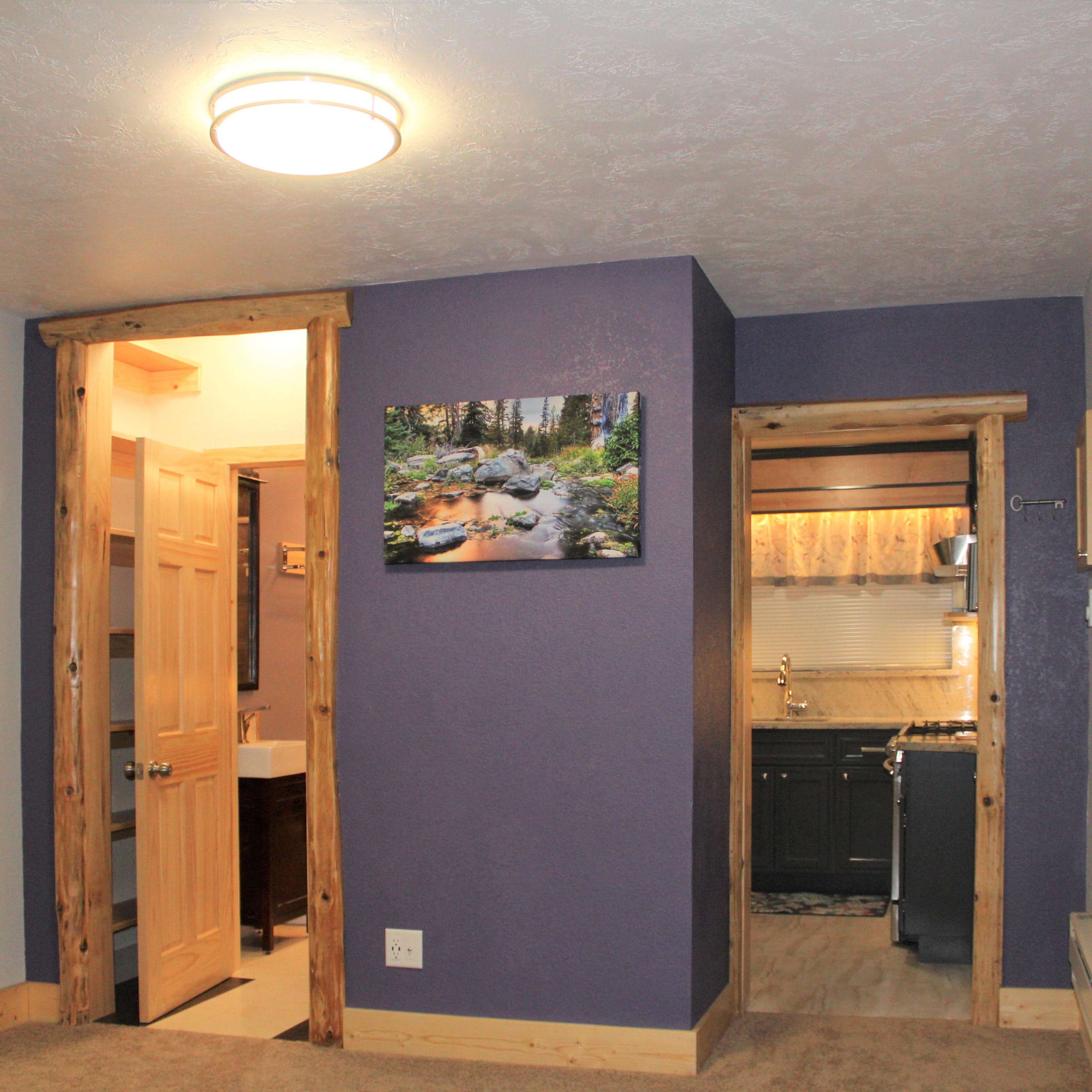 A room with a purple wall, wooden accents, and a doorway leading to a bathroom and kitchen area, with a landscape painting hanging on the wall.