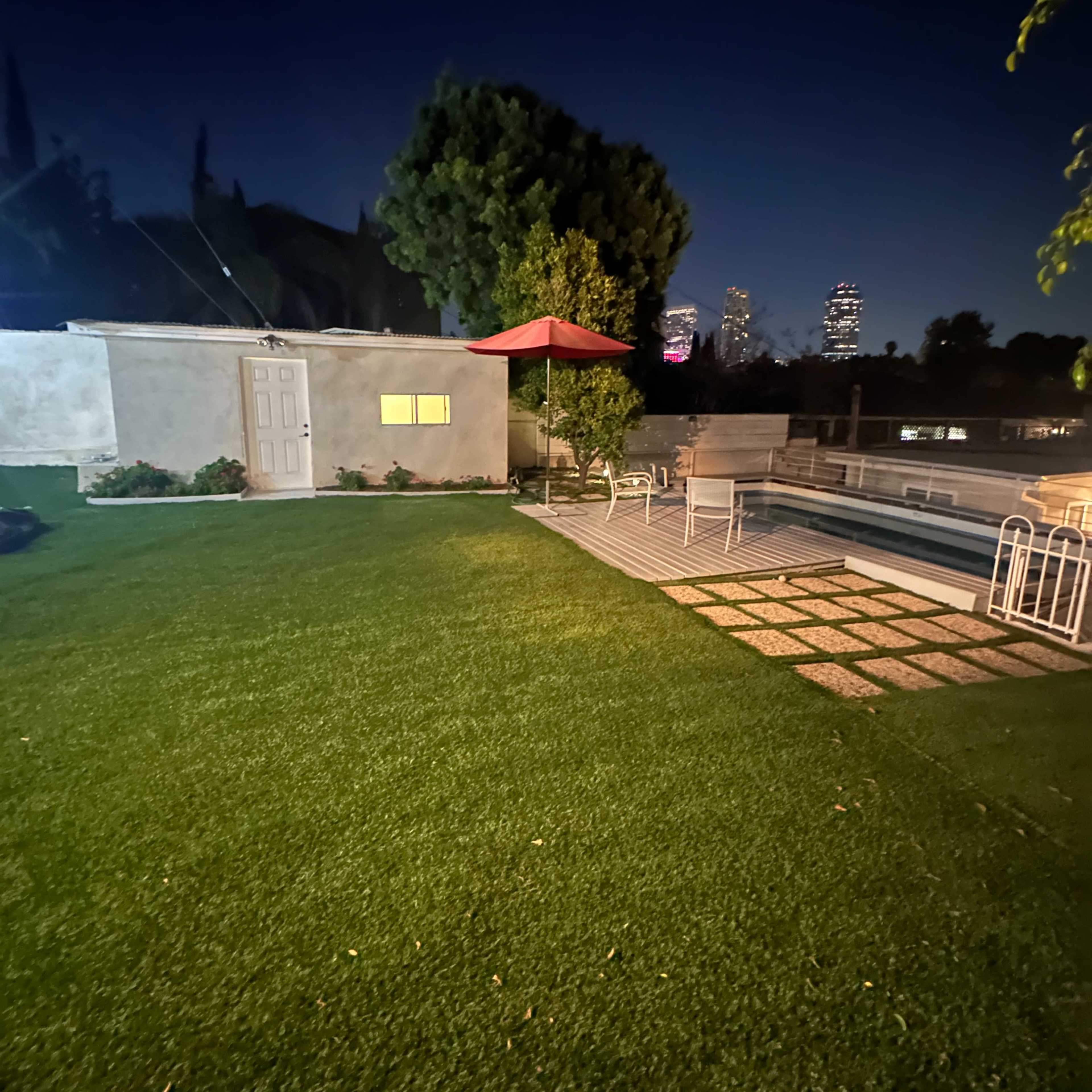 A well-manicured backyard at night, featuring a small shed, a red umbrella over a seating area, and a pool beside a stone patio.