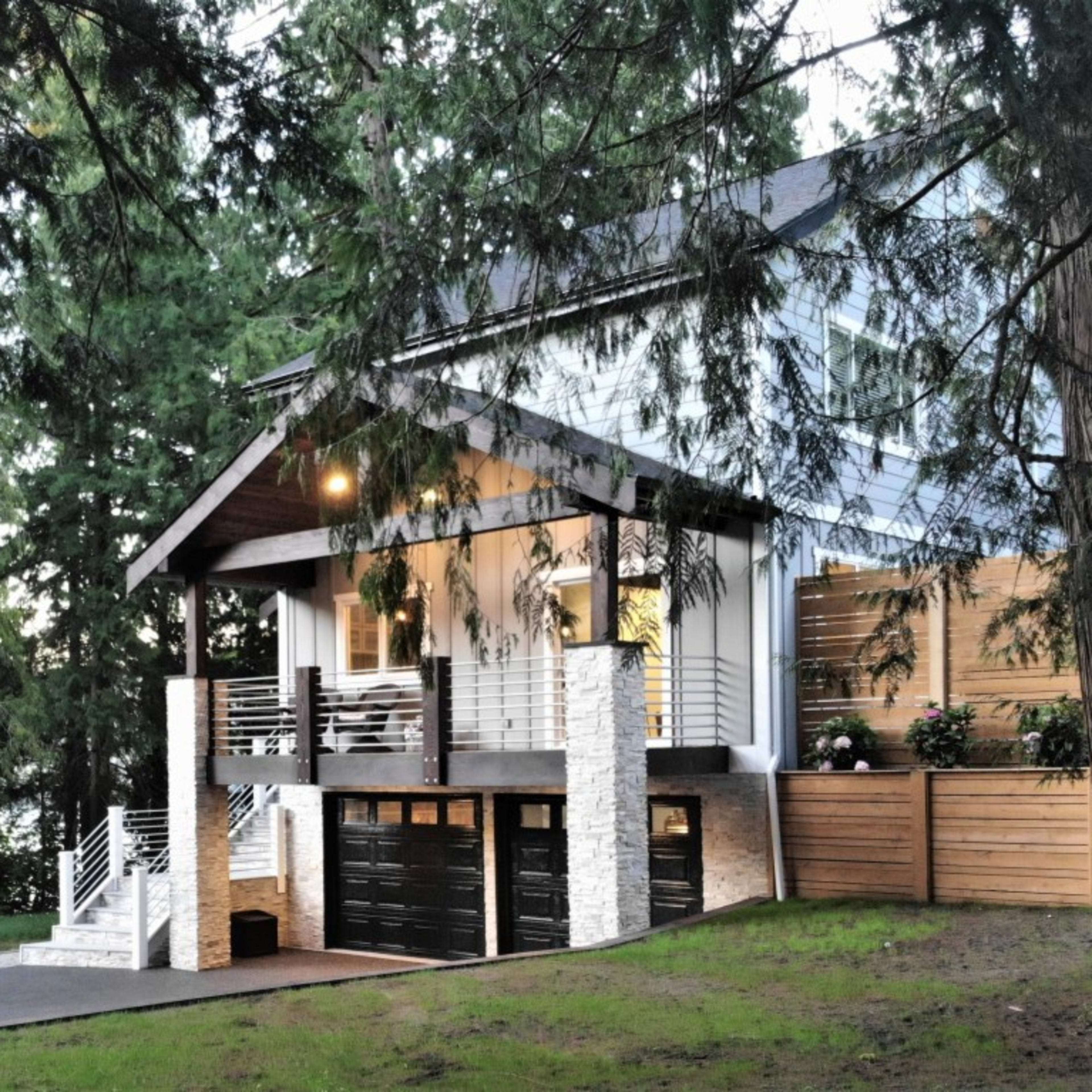 A modern house with a covered porch and two garage doors is surrounded by tall trees.