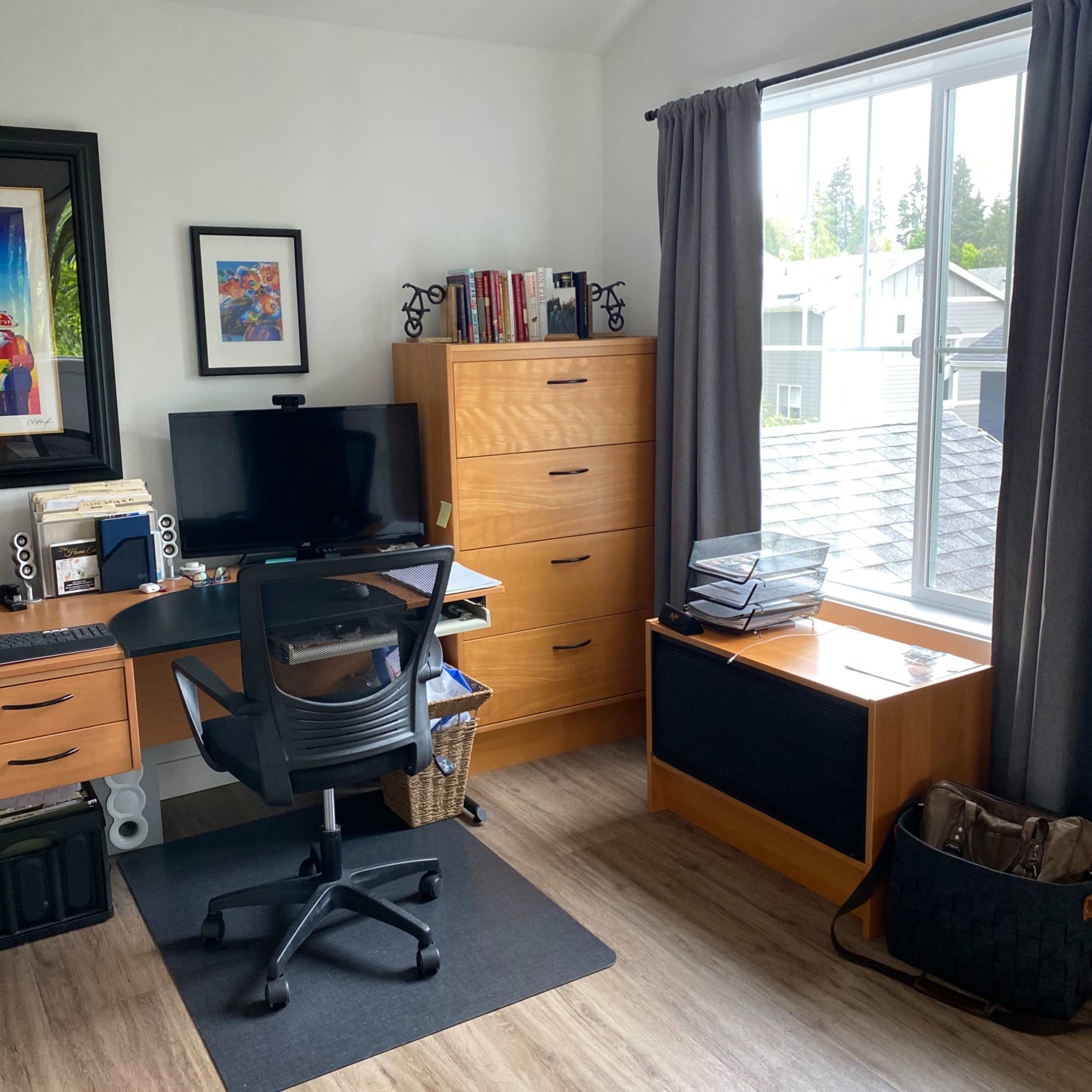 A modern home office features a black desk and chair, a monitor, a tall wooden dresser, and a large window with gray curtains.