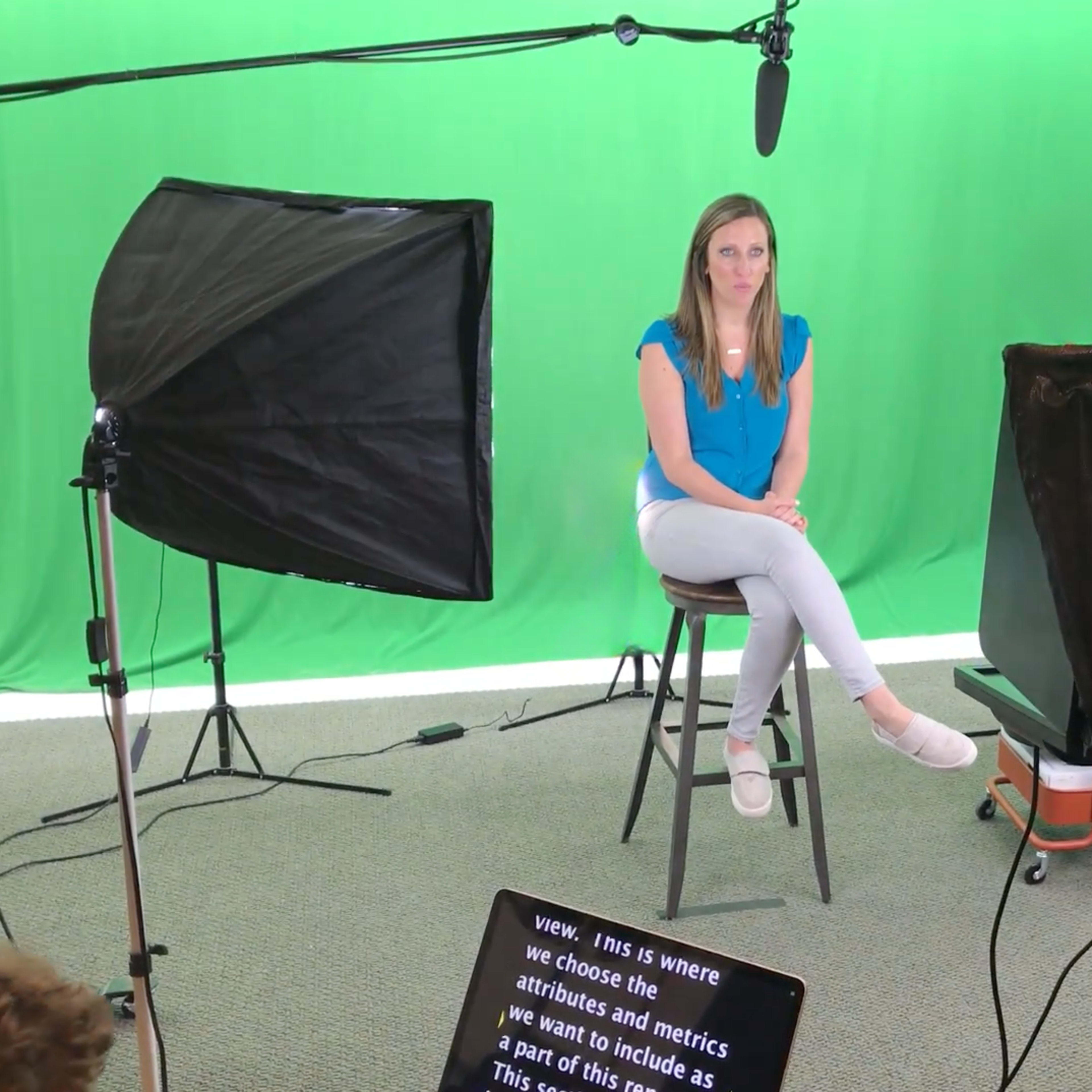 A woman sits on a stool in front of a green screen, surrounded by lighting equipment and a camera setup.
