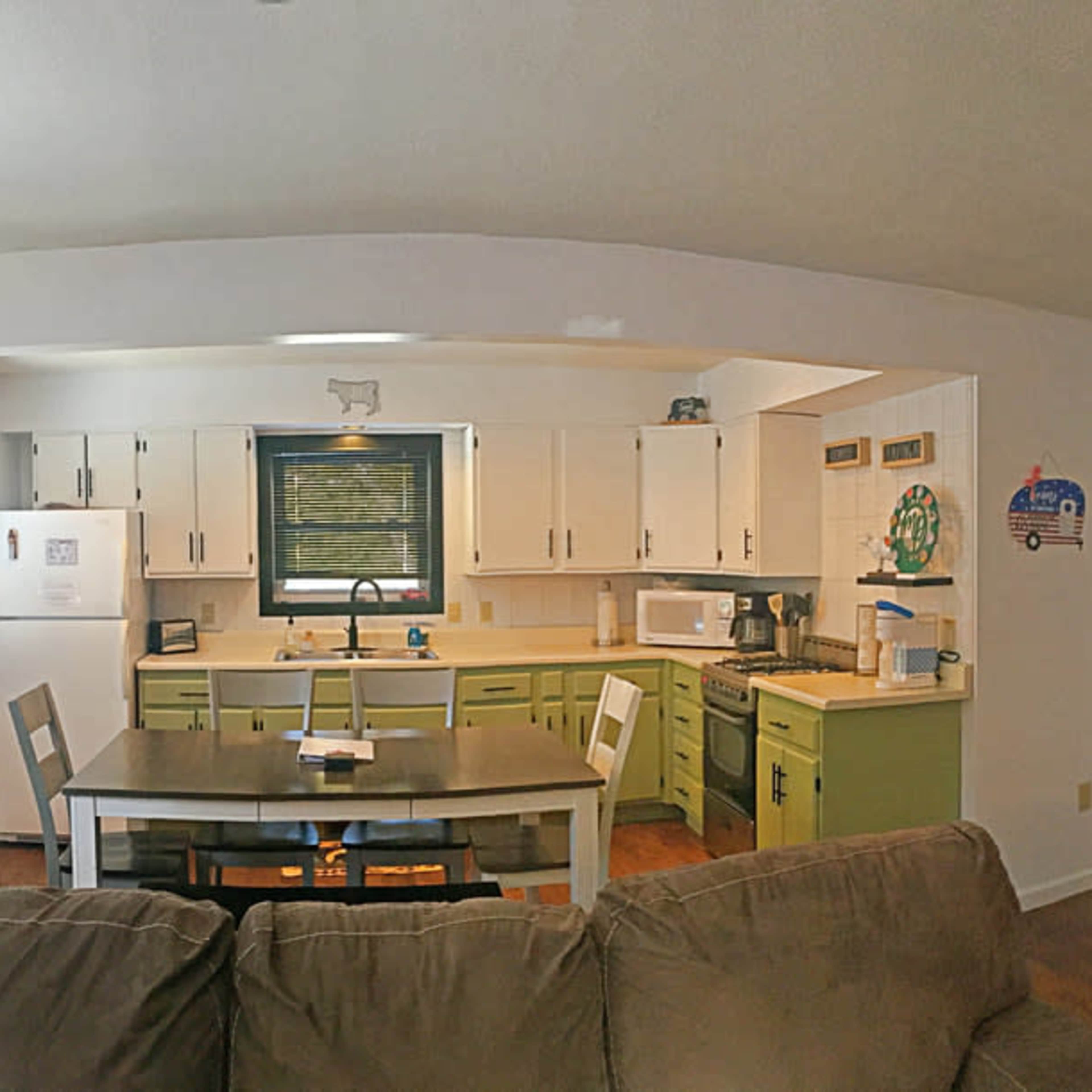 A bright, open-concept kitchen and dining area with green cabinetry, a white refrigerator, and a staircase in the background.