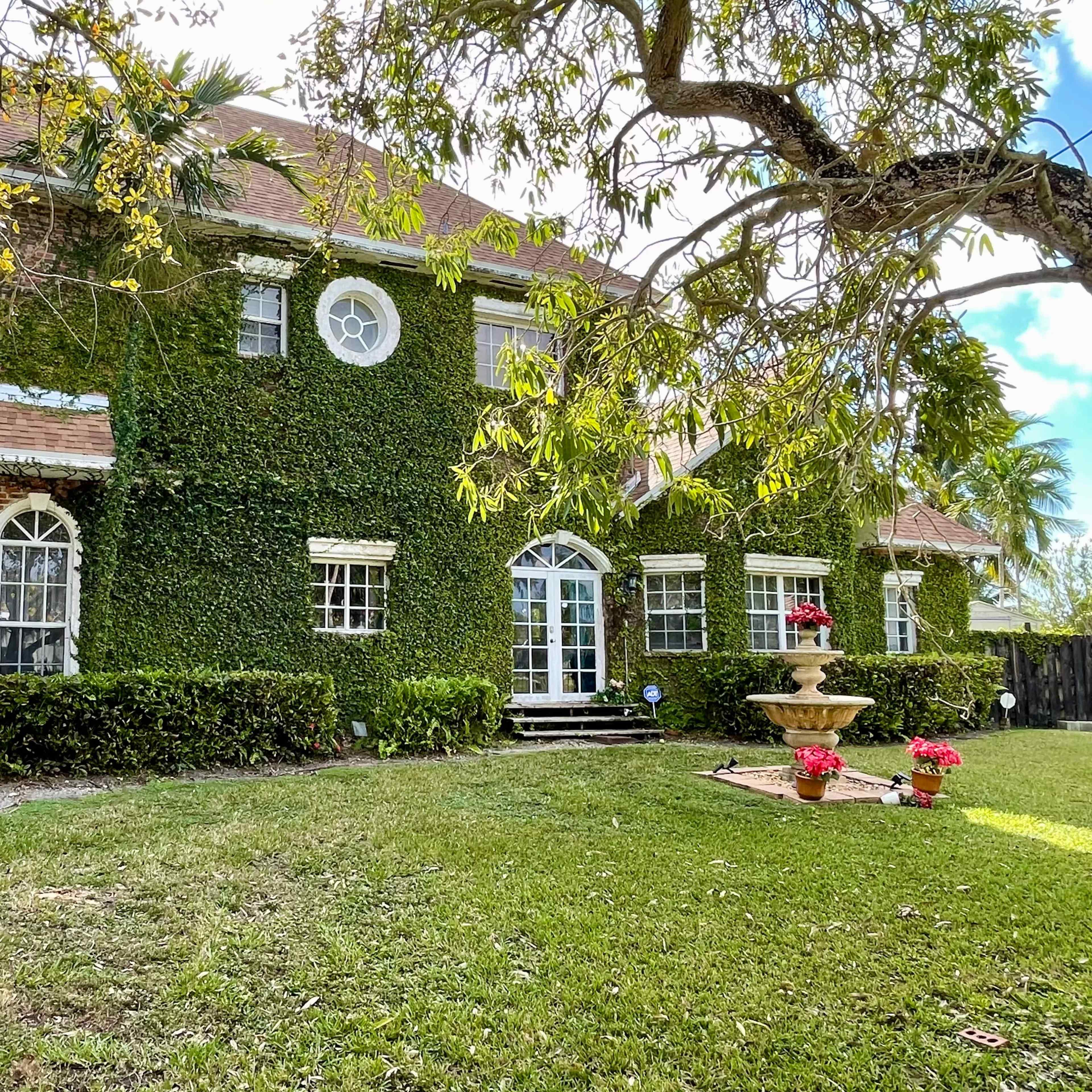 A two-story brick house covered in ivy is surrounded by a well-maintained lawn and features a central fountain with potted flowers in the front yard.