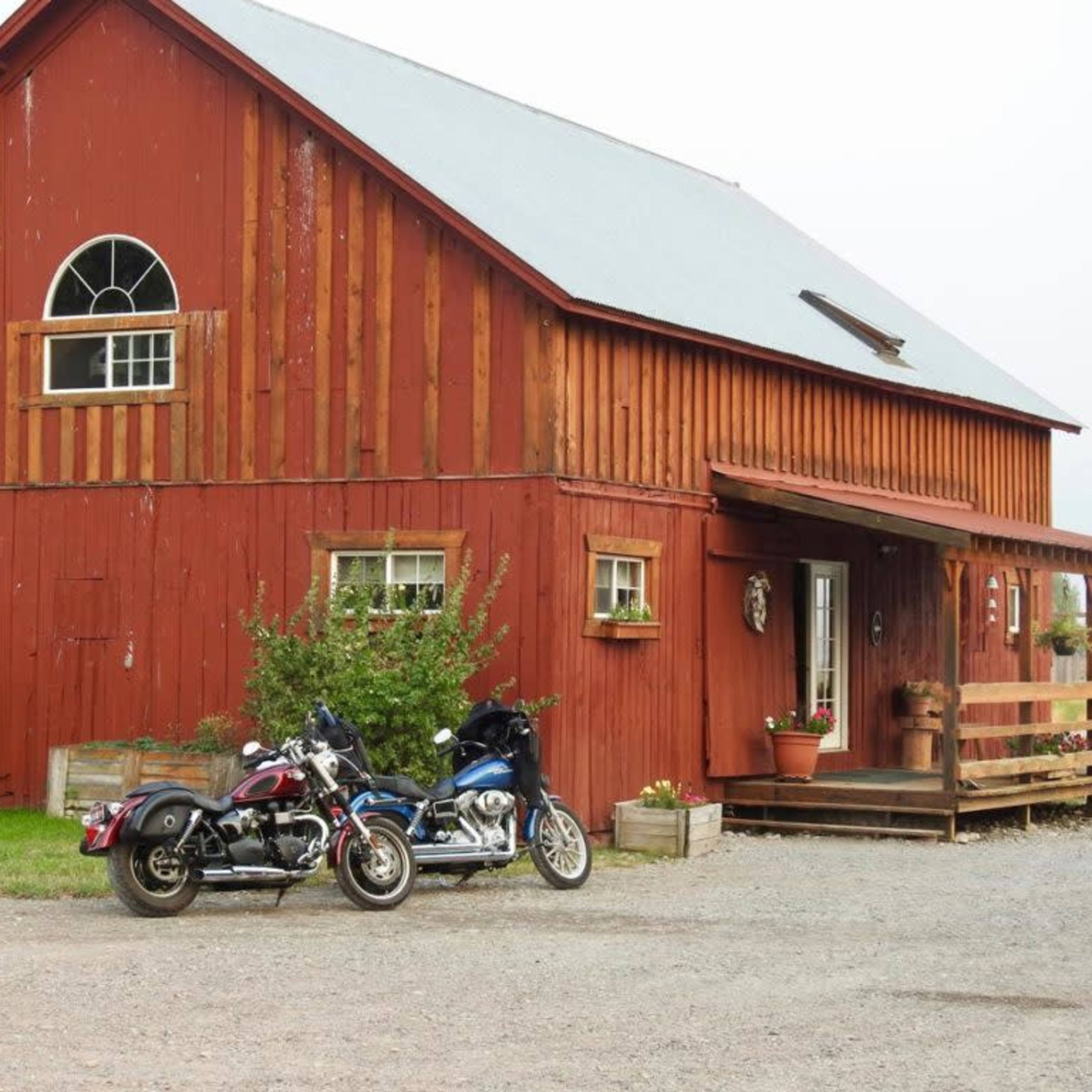 Two motorcycles are parked in front of a red barn with a porch and several windows.