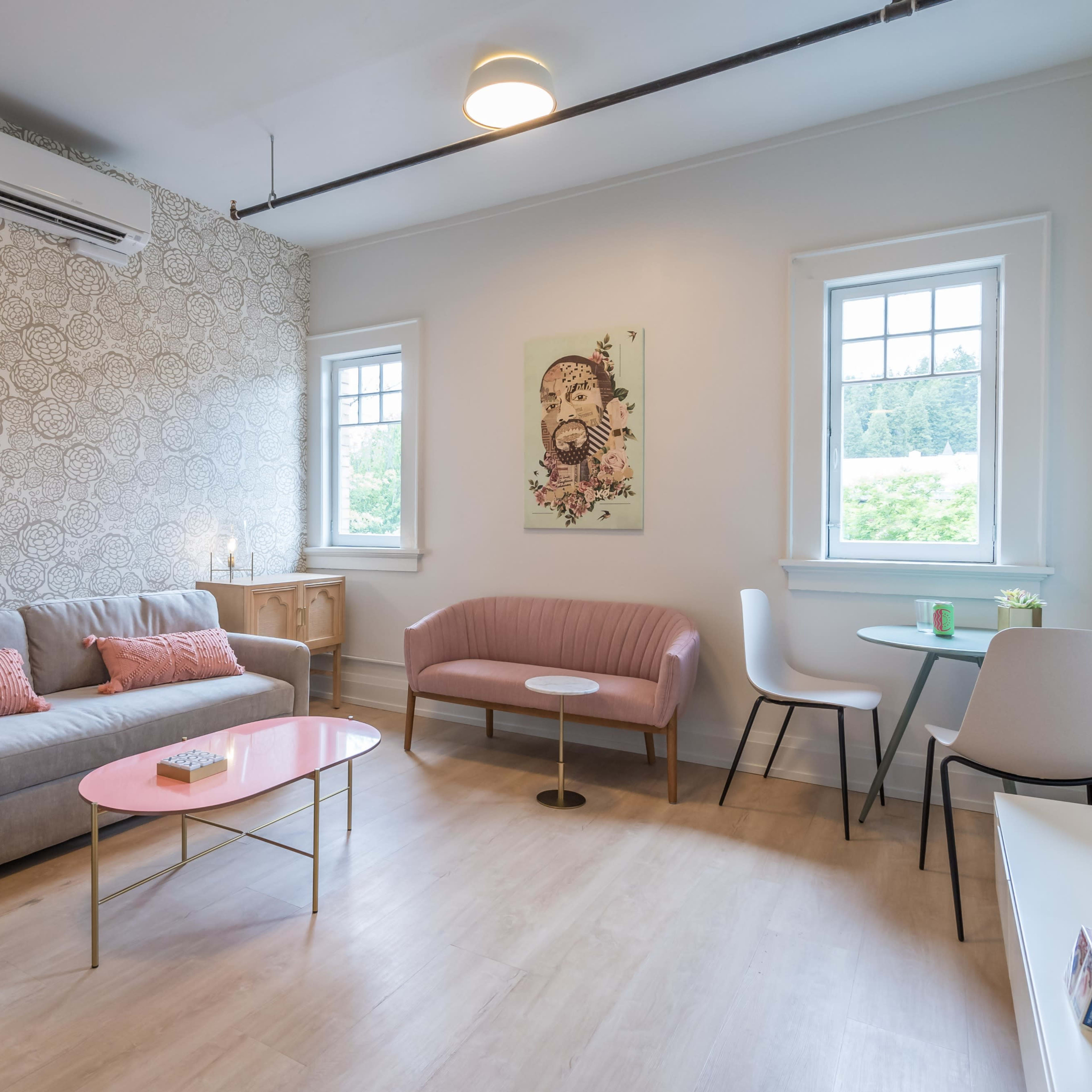 A living room with a gray sofa, a pink chair, a light pink coffee table, and two windows overlooking greenery.